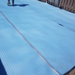 Kamil Perczak - Niebieska pianka izolacyjna z siatką, częściowo rozłożona na poddaszu, widoczne łączenia i fragment drewnianego płotu w tle.