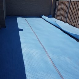 Kamil Perczak - Niebieska mata izolacyjna z siatką ułożona na podłożu, częściowo rozwinięta, widoczna ściana z cegły i drewniany płot w tle.