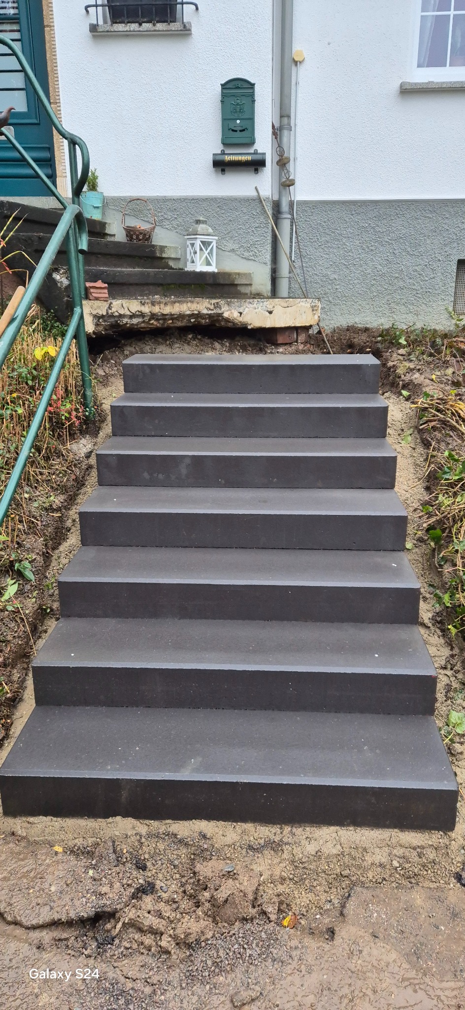 Nowe, ciemnoszare, betonowe schody zewnętrzne prowadzące do domu z zieloną skrzynką pocztową, widoczne pozostałości po starych schodach i ziemia po bokach.
