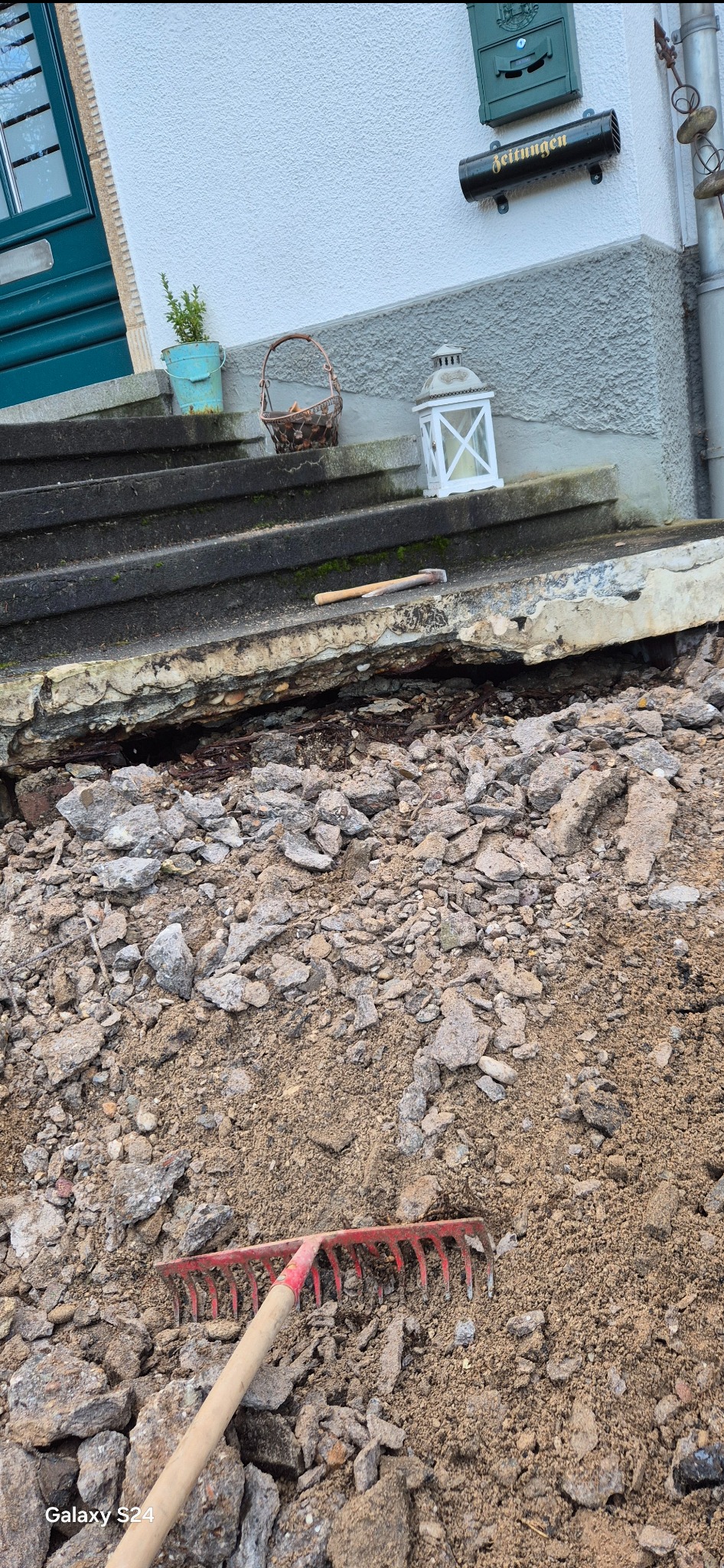 Uszkodzone betonowe schody wejściowe do domu w trakcie remontu, gruz i ziemia na pierwszym planie, widoczny młotek i grabie.