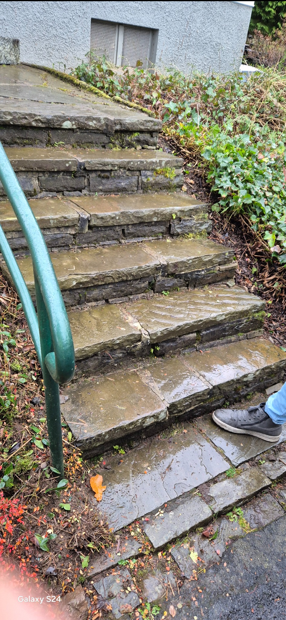 Mokre kamienne schody zewnętrzne z zieloną metalową poręczą, częściowo widoczna stopa w szarym bucie i niebieskich dżinsach.