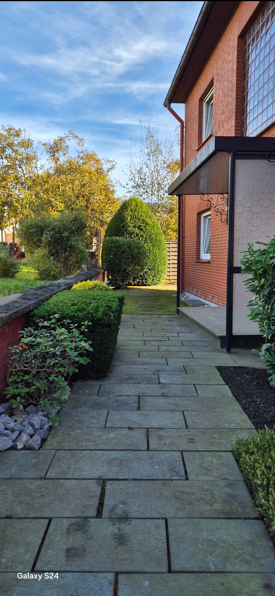 Kamienna ścieżka prowadząca do wejścia domu z czerwonej cegły, otoczona zadbanym ogrodem z równo przyciętymi krzewami i drzewami w jesiennych barwach.