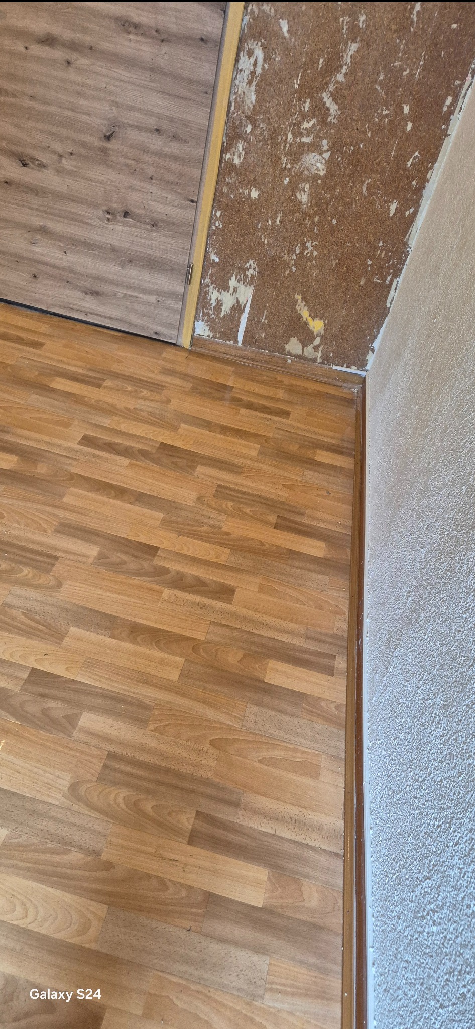 Fragment pokoju w trakcie prac: panele podłogowe ułożone, ściana z resztkami tapety i surowym tynkiem, widoczna listwa przypodłogowa.