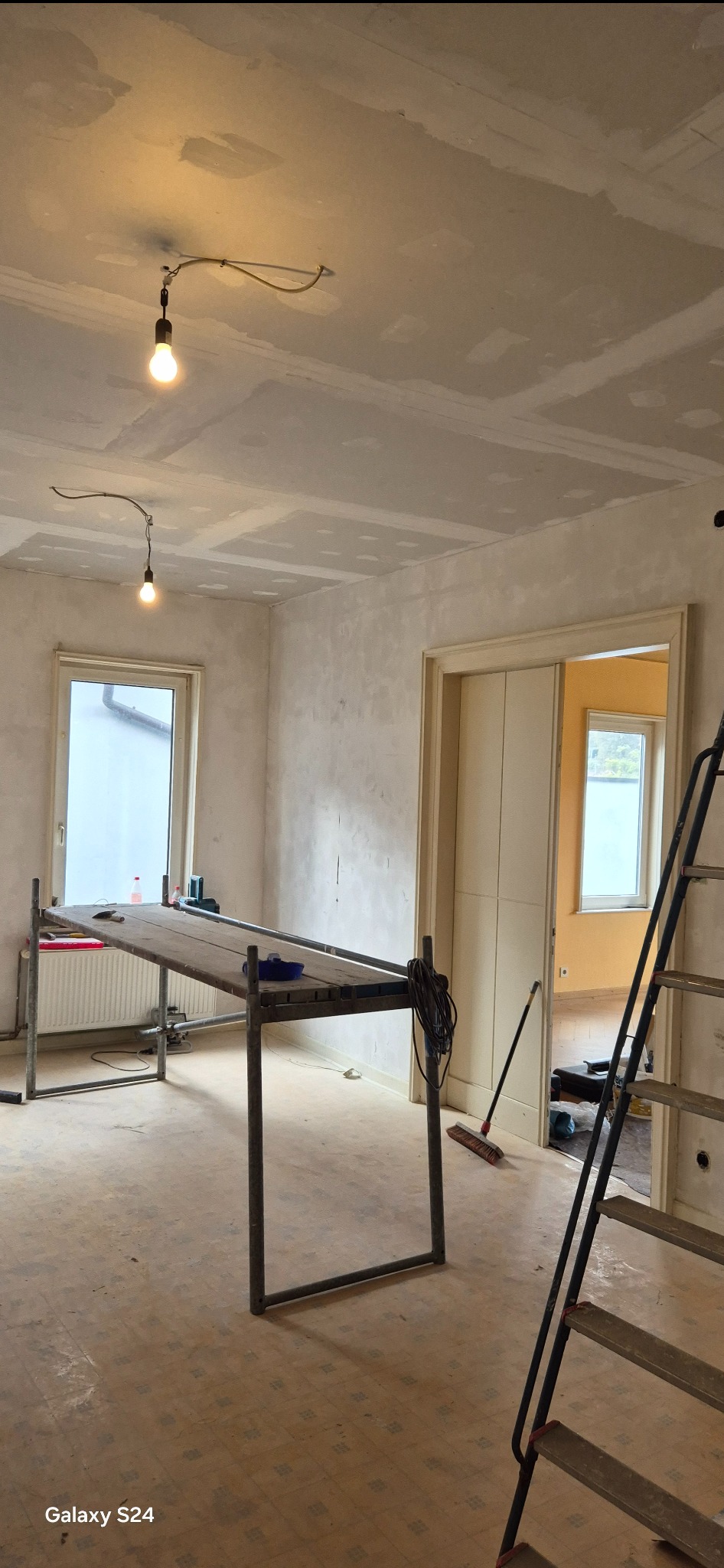 Wnętrze pokoju w trakcie prac wykończeniowych: surowe ściany i sufit pokryte płytami gipsowo-kartonowymi, prowizoryczne oświetlenie, metalowy stół roboczy z narzędziami, drabina i widoczne ślady...