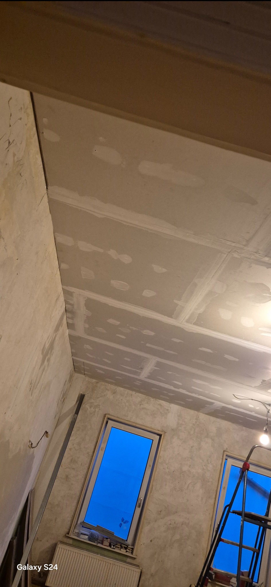Poddasze w trakcie ocieplania: widoczne płyty gipsowo-kartonowe na suficie, szpachlowane łączenia, surowe ściany i okna z niebieskim światłem dziennym. Rozstawiona drabina w rogu pokoju.