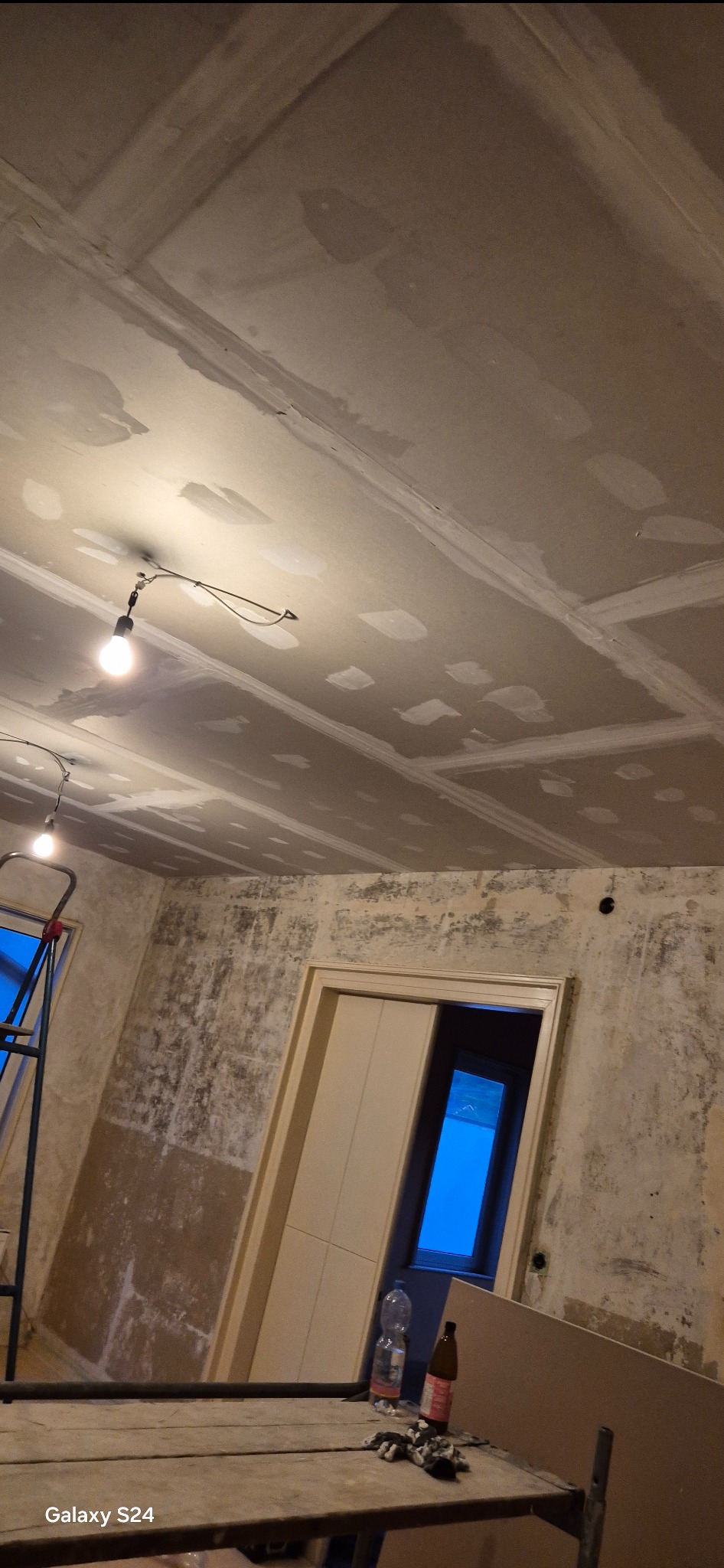 Pokój w trakcie prac przygotowawczych do malowania: zagruntowany sufit z widocznymi łączeniami płyt gipsowo-kartonowych, ściany z pozostałościami starej farby, okno z widokiem na zewnątrz, butelki...