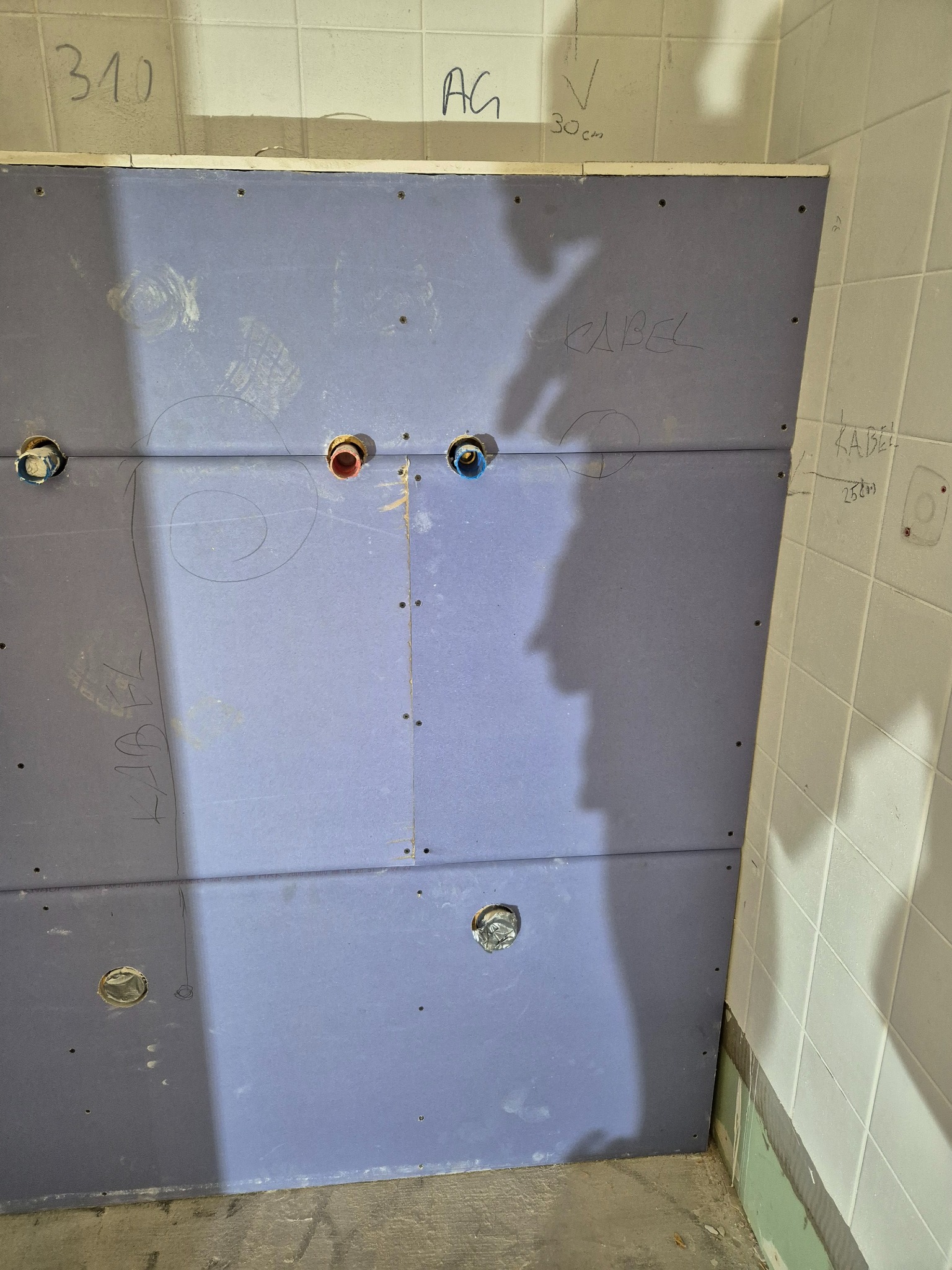 Ściana z płyt gipsowo-kartonowych w łazience, z widocznymi otworami na instalacje wodne, tymczasowe notatki i oznaczenia na ścianie, ukazująca etap przygotowawczy do wykończenia.