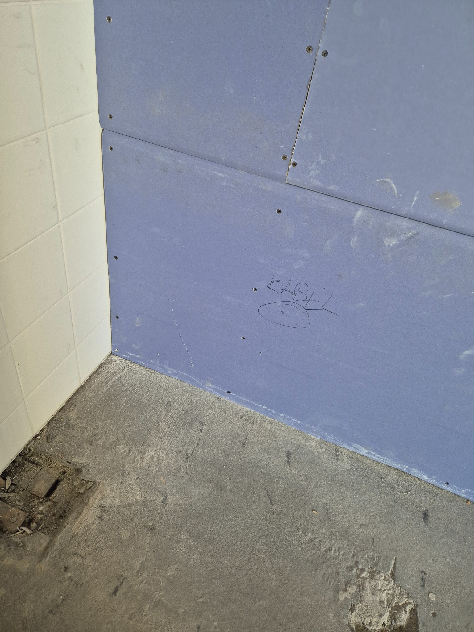 Narożnik łazienki w trakcie prac: białe płytki ceramiczne, niebieska płyta gipsowo-kartonowa z napisem 'KABEL' i surowy beton na podłodze z widocznymi uszkodzeniami.
