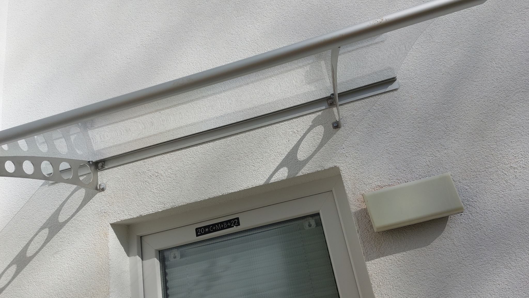 Szklany daszek nad oknem z widocznym napisem '20*C+M+B+22' na ramie okiennej i skrzynką na listy obok, zamontowany na elewacji budynku o szorstkiej fakturze.