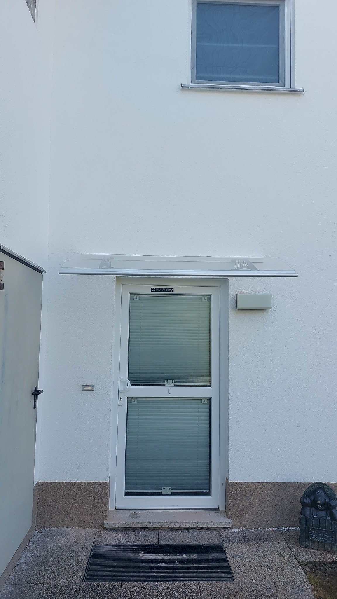 Białe drzwi wejściowe z żaluzjami wewnętrznymi i przezroczystym zadaszeniem, numer domu 20*C+M+B+22, posąg psa po prawej stronie, biała elewacja budynku z małym oknem u góry.