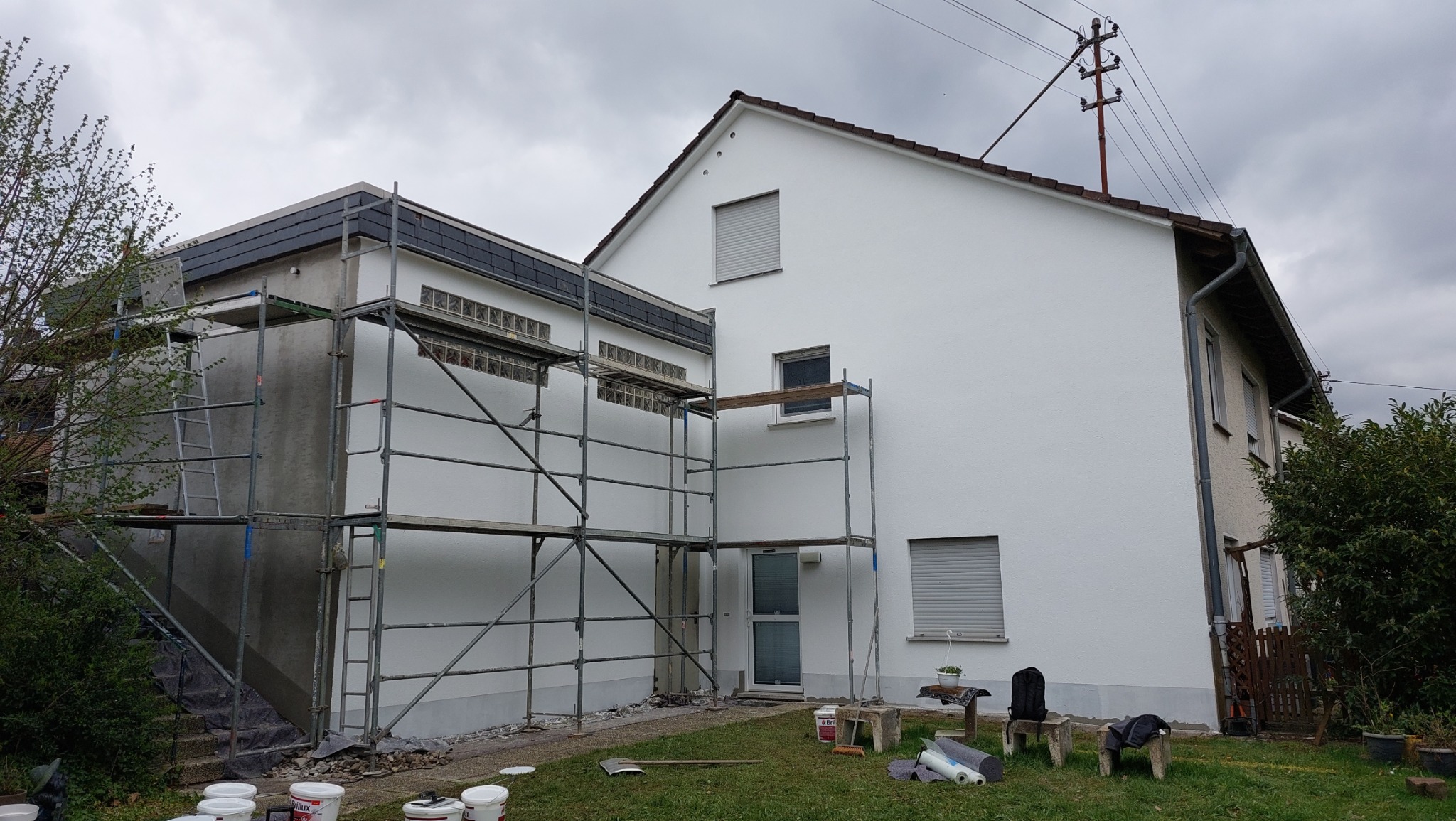 Dom jednorodzinny w trakcie malowania elewacji, widoczne rusztowania, puste puszki po farbie na trawniku i fragmenty ocieplonego poddasza z szarymi płytkami.