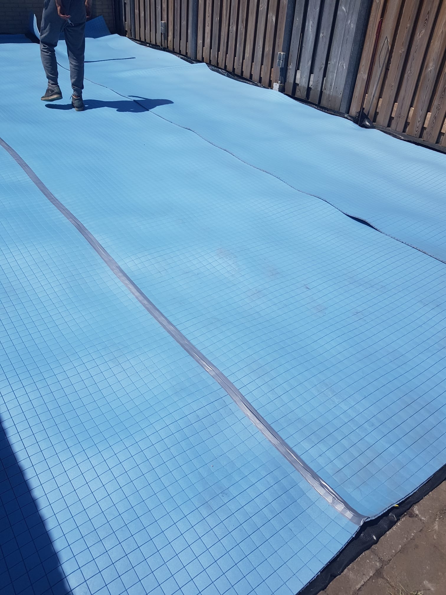 Niebieska pianka izolacyjna z siatką, częściowo rozłożona na poddaszu, widoczne łączenia i fragment drewnianego płotu w tle.