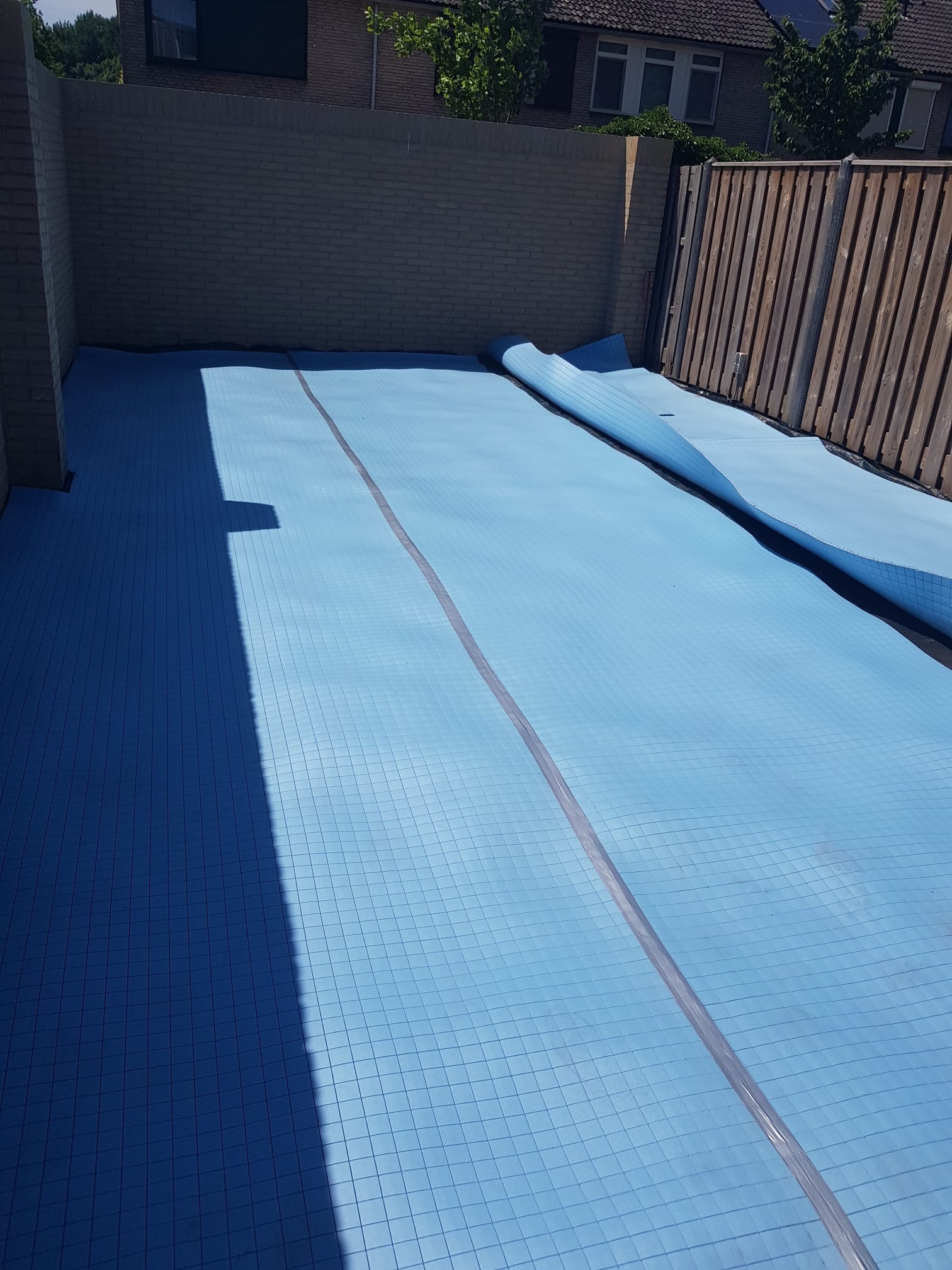 Niebieska mata izolacyjna z siatką ułożona na podłożu, częściowo rozwinięta, widoczna ściana z cegły i drewniany płot w tle.
