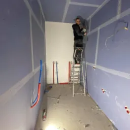 Pracownik na drabinie szpachluje łączenia płyt gipsowo-kartonowych na suficie w pomieszczeniu z wyprowadzonymi instalacjami wodnymi i elektrycznymi, widoczne narzędzia i resztki materiałów...