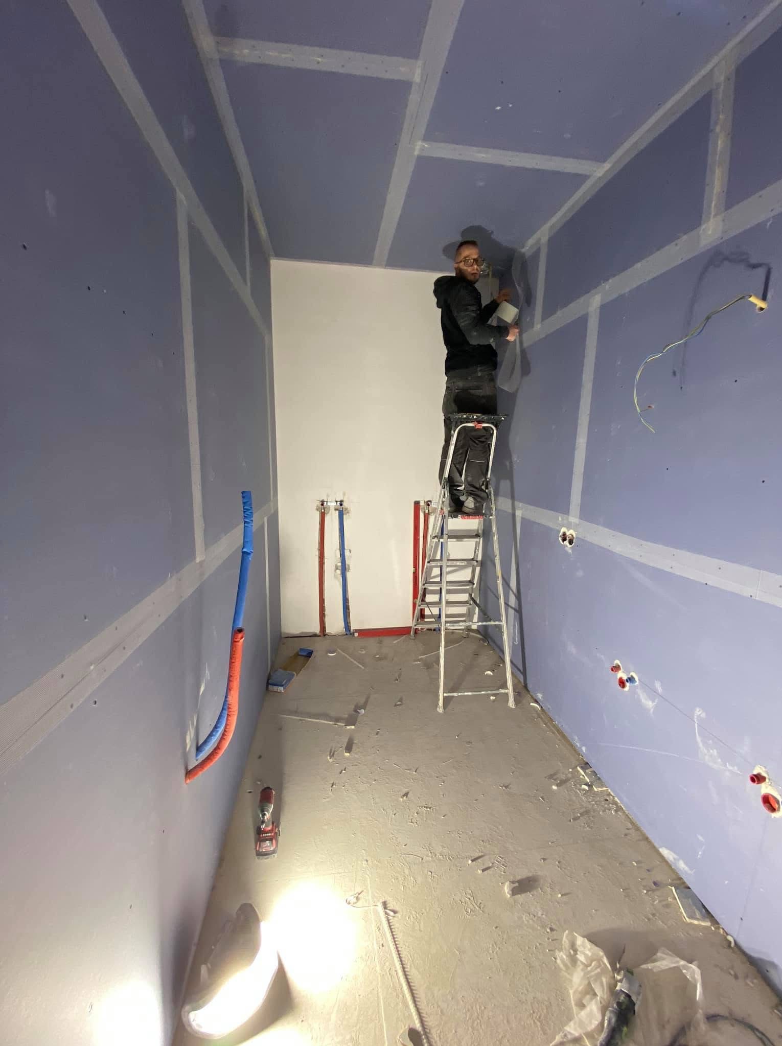 Pracownik na drabinie szpachluje łączenia płyt gipsowo-kartonowych na suficie w pomieszczeniu z wyprowadzonymi instalacjami wodnymi i elektrycznymi, widoczne narzędzia i resztki materiałów...