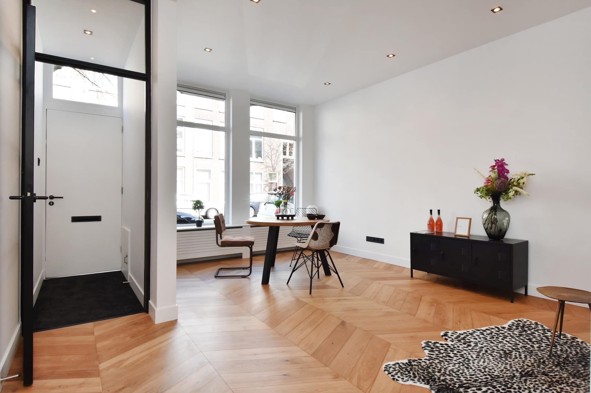 Jasne wnętrze z parkietem ułożonym w jodełkę, czarnym kredensem z dekoracjami, okrągłym stołem przy oknie i lustrem w czarnej ramie przy wejściu.