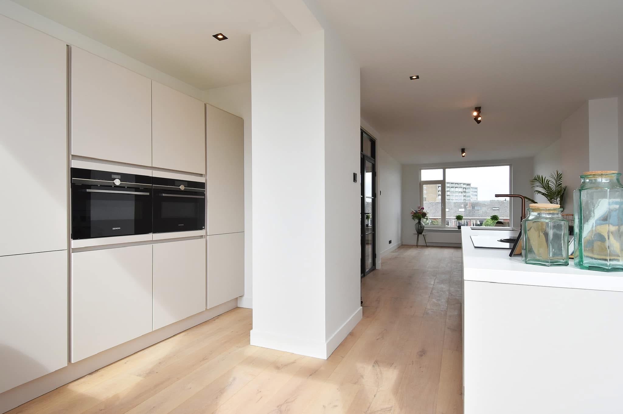 Jasne, minimalistyczne wnętrze kuchni z drewnianą podłogą, zabudową kuchenną z dwoma piekarnikami Siemens i widokiem na salon oraz okno z widokiem na miasto.