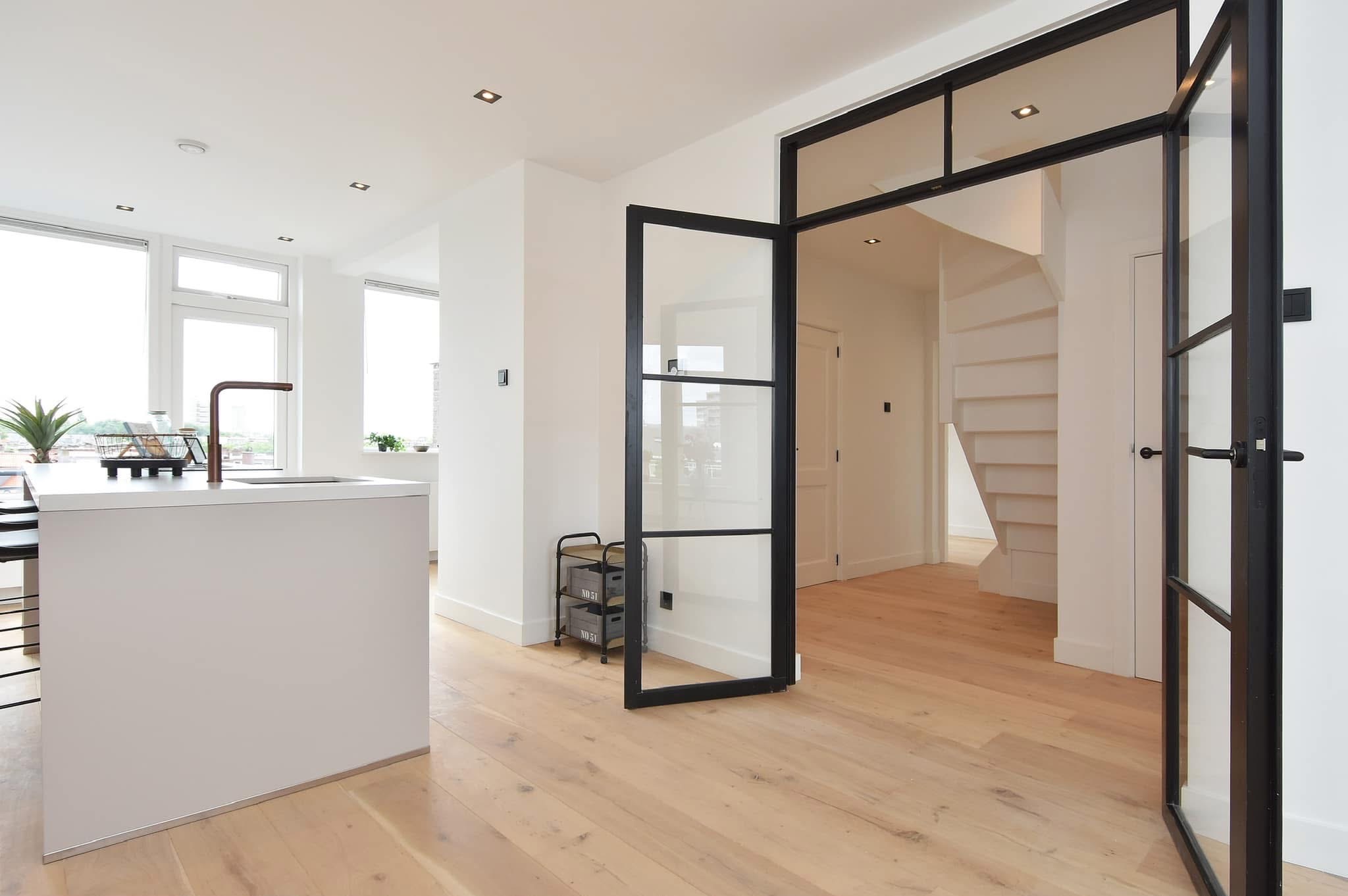 Jasne wnętrze mieszkania z drewnianą podłogą, białymi ścianami i czarną, przeszkloną ścianką działową z drzwiami, widok na białe schody i wyspę kuchenną z miedzianą baterią.