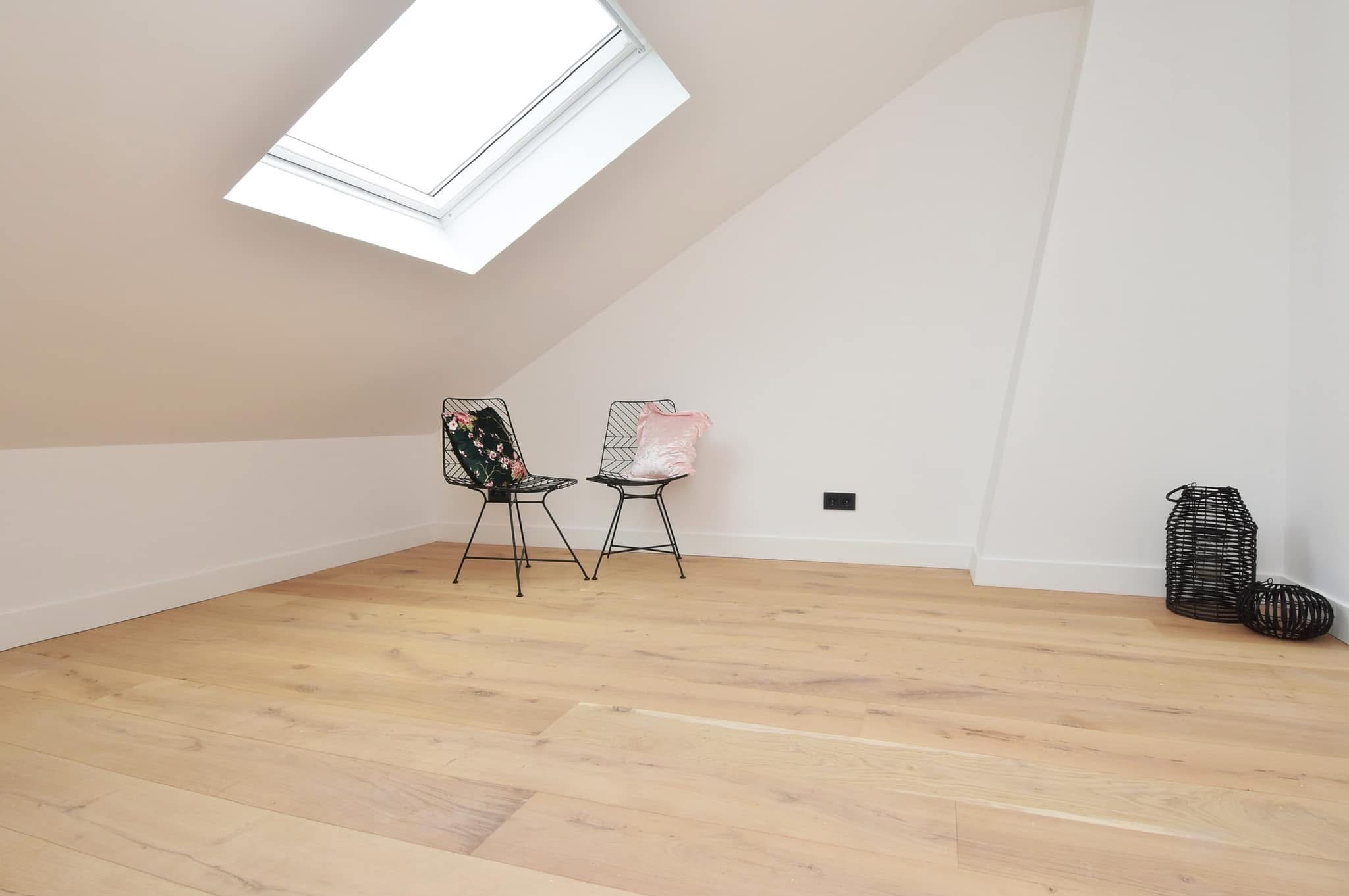 Jasne poddasze z oknem dachowym, parkietem z jasnego drewna, dwoma metalowymi krzesłami z poduszkami i dekoracyjnymi lampionami w rogu.
