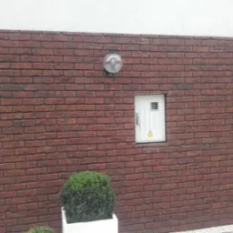 Czerwona ściana z cegły klinkierowej z oświetleniem kulistym, białą skrzynką elektryczną z symbolem ostrzegawczym i rurą spustową, uzupełniona o kulisty krzew w białej donicy.