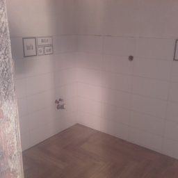 Jurek-PROF - Wnętrze łazienki w trakcie wykańczania: białe płytki na ścianach z dekoracyjnymi napisami w ramkach, drewnopodobne panele ułożone w jodełkę na podłodze, widoczne wystające rury instalacji wodnej...