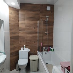 Jurek-PROF - Ukończona łazienka z brązowymi płytkami imitującymi drewno na ścianie, białą armaturą, pralką i wanną za szklanym panelem.