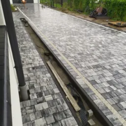 Układanie szarej kostki brukowej na podjeździe, widoczny liniowy system odprowadzania wody z piaskiem i elementami montażowymi, w tle garaż i osoby przy betoniarce.