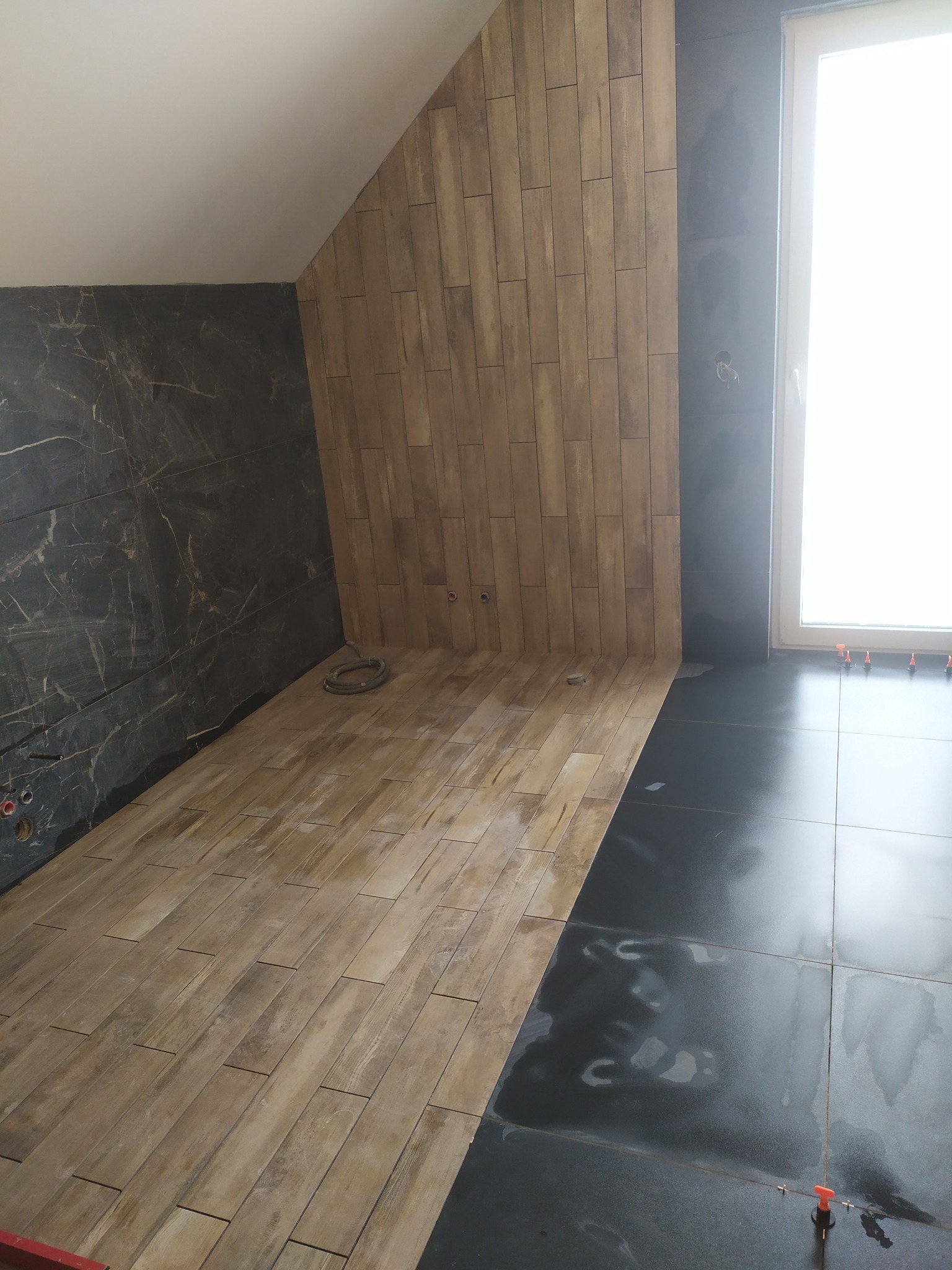 Wnętrze łazienki w trakcie wykańczania, z częściowo ułożonymi płytkami imitującymi drewno na podłodze i ścianie pod skosem, oraz ciemnymi płytkami w strefie przy oknie, z widocznymi elementami...