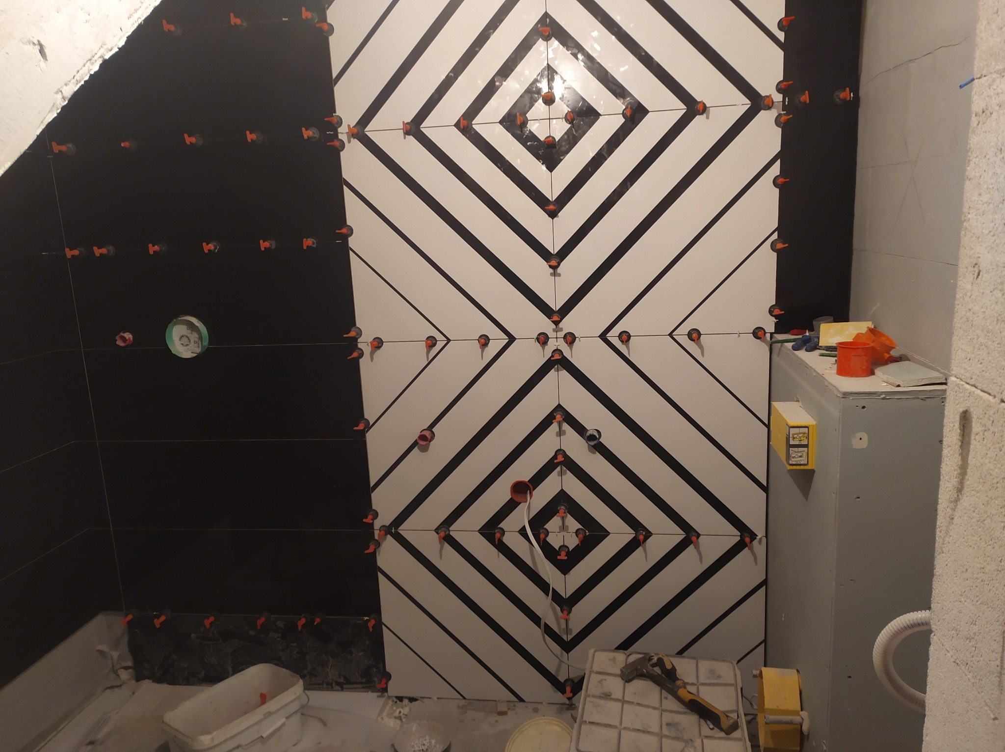 Ściana łazienki w trakcie układania płytek w geometryczny wzór z użyciem klipsów poziomujących, widoczne narzędzia i elementy instalacji.