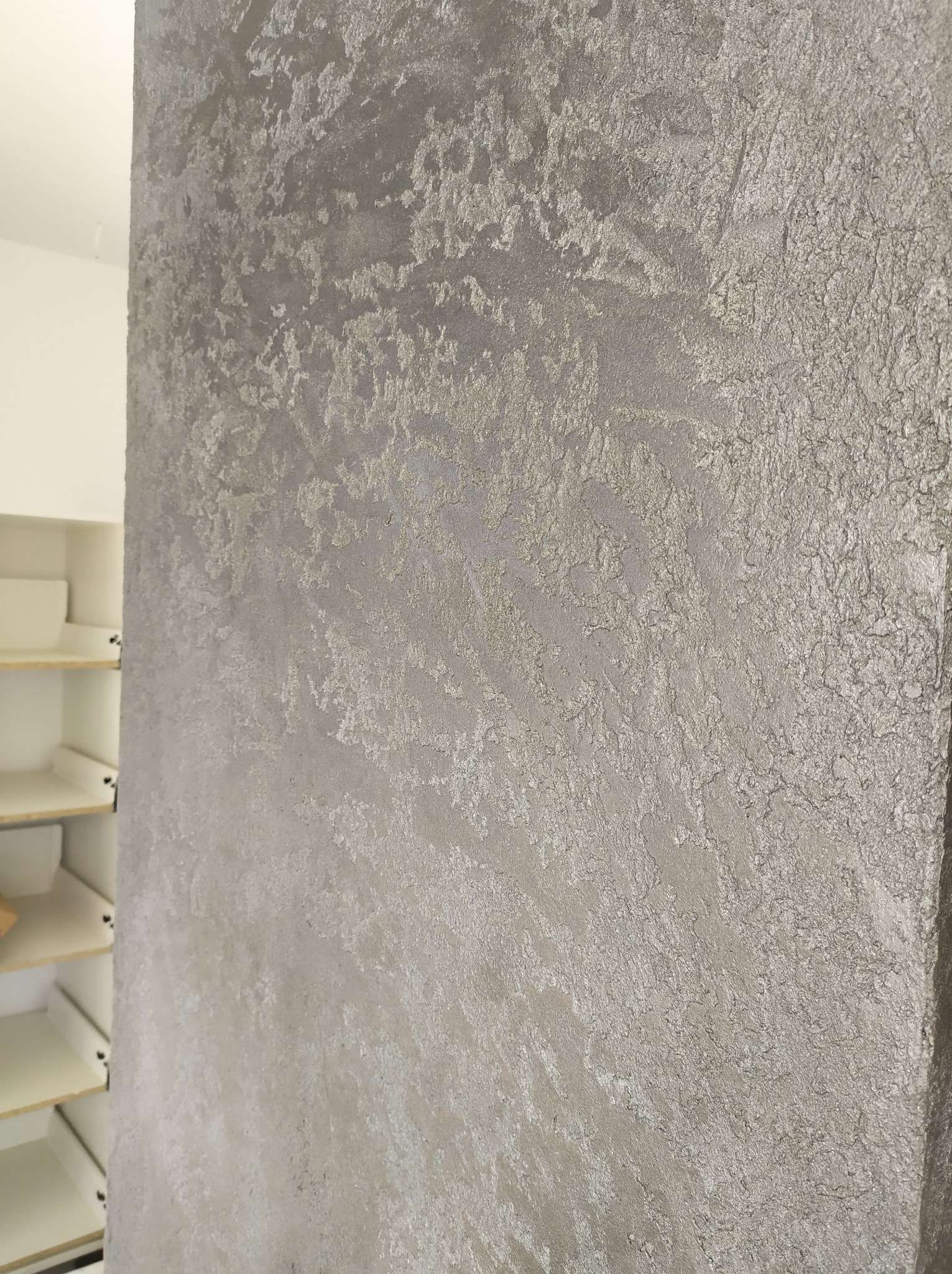Teksturowana ściana w odcieniach szarości z efektem metalicznego połysku, widoczna z bliska, z fragmentem białych półek w tle.