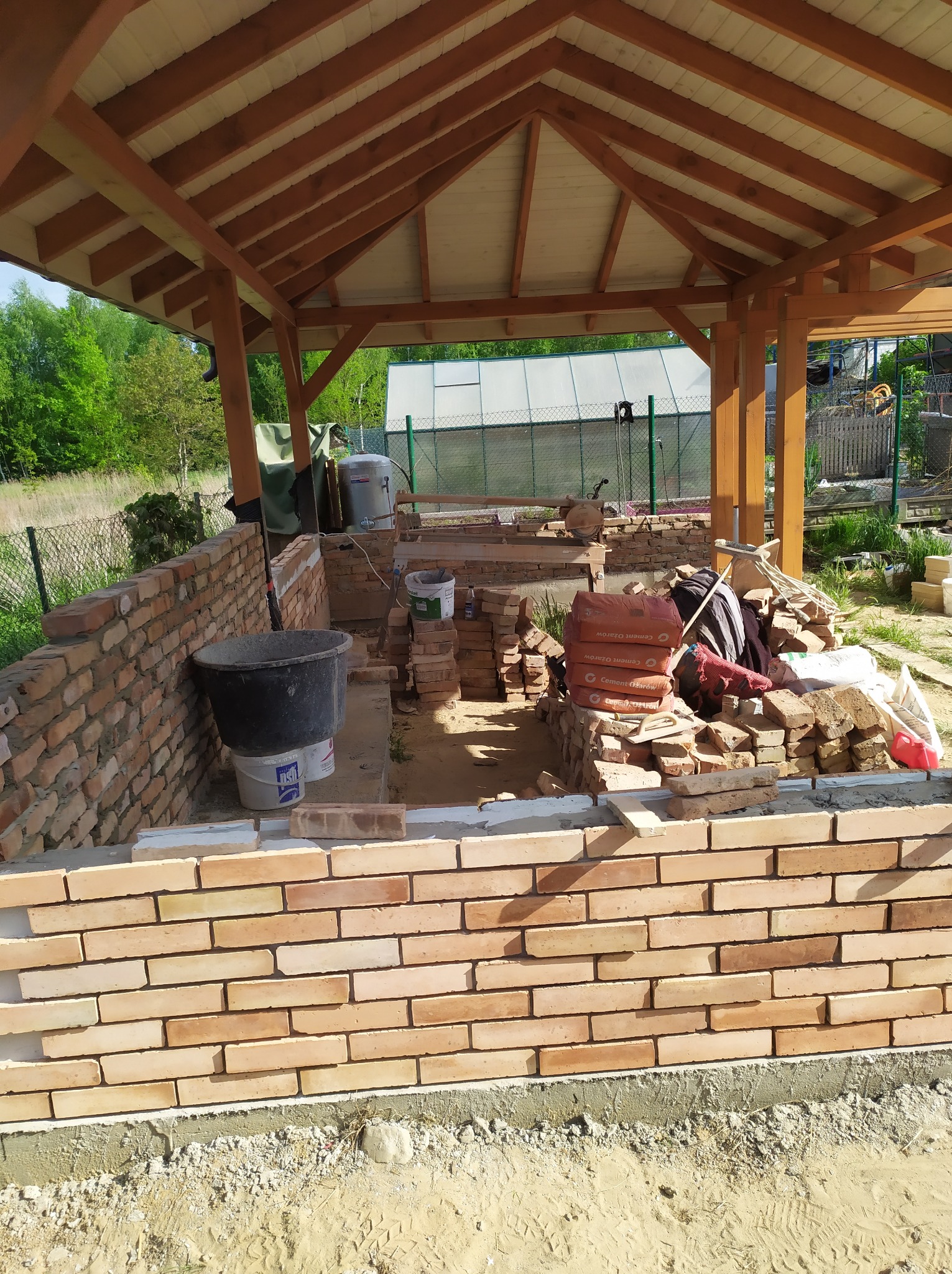 Wznoszenie ścian z cegły klinkierowej pod drewnianą konstrukcją zadaszenia, widoczne worki cementu i narzędzia budowlane, etap budowy altany lub grilla.