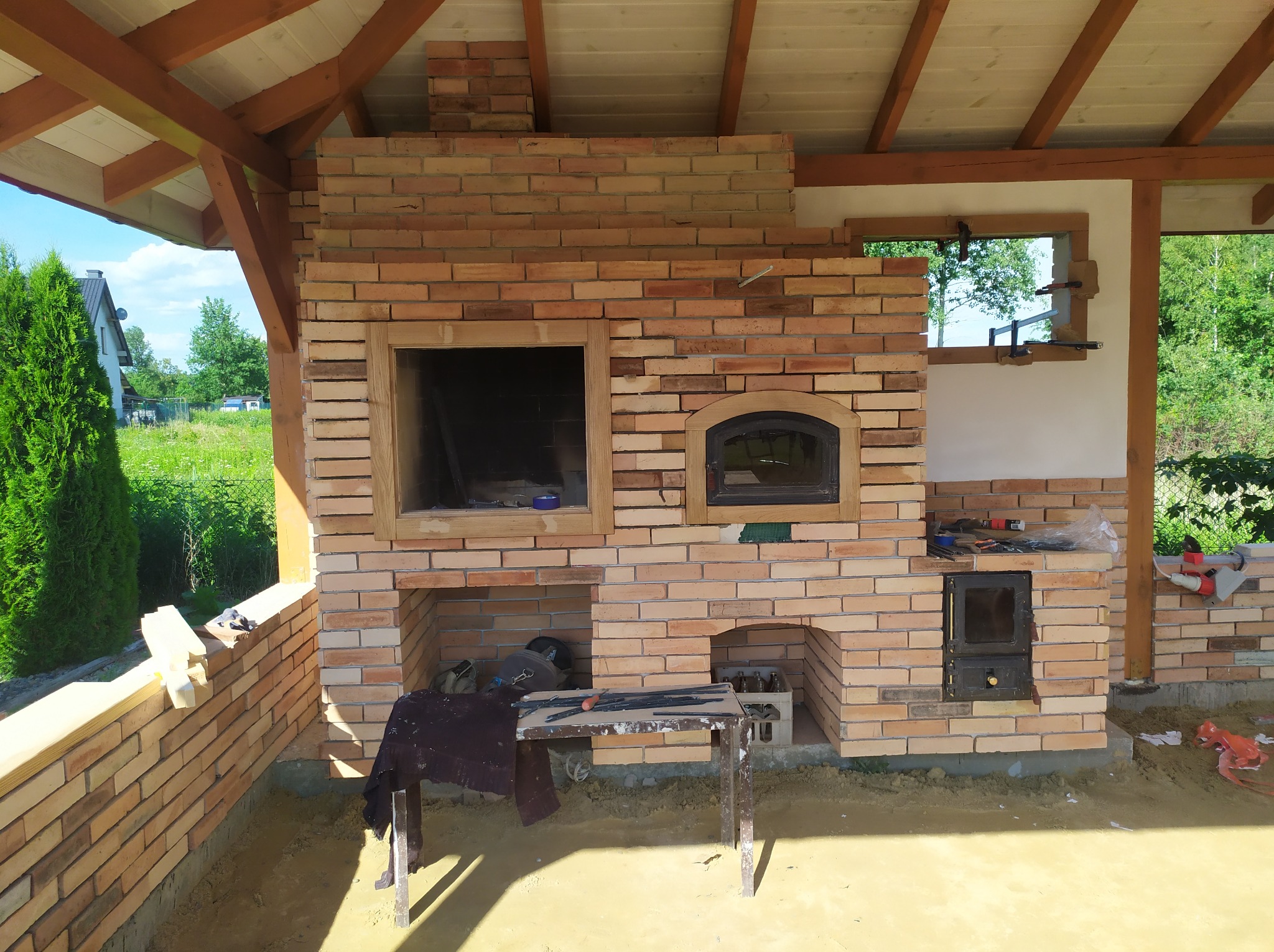 Murowany piec zewnętrzny z cegły w trakcie budowy, widoczne elementy konstrukcyjne, stół roboczy z narzędziami i niezagospodarowany teren wokół.