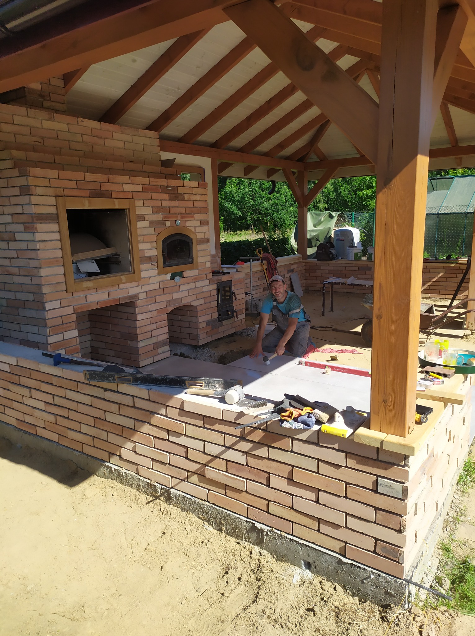 Uśmiechnięty mężczyzna w trakcie układania jasnych płytek na ceglanym podwyższeniu, prawdopodobnie przygotowywanym pod budowę pieca w altanie. Widoczne narzędzia murarskie, poziomica i ceglany piec...