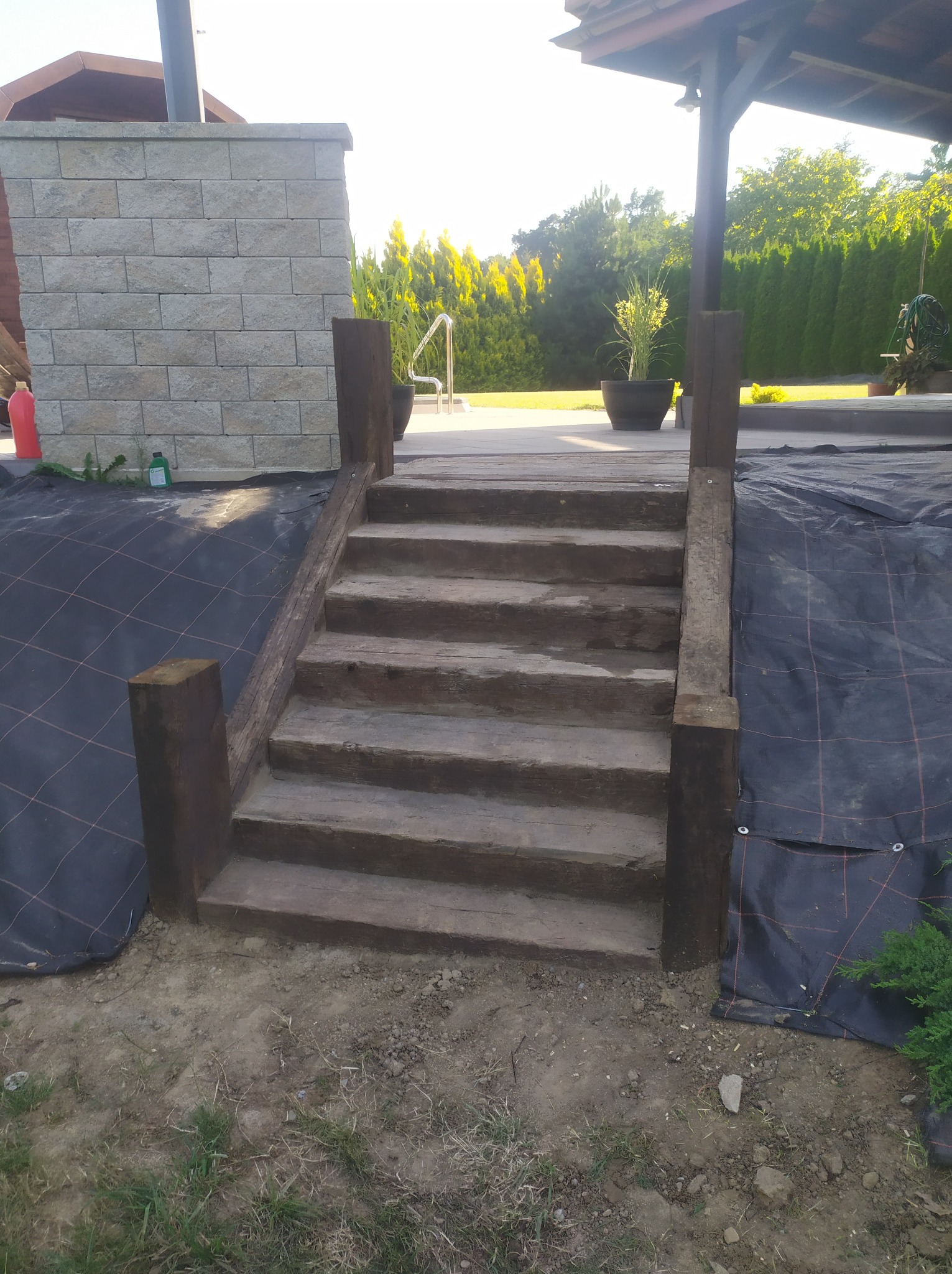 Drewniane schody prowadzące na taras, z widoczną czarną tkaniną agrowłókninową w tle i elementami krajobrazu.