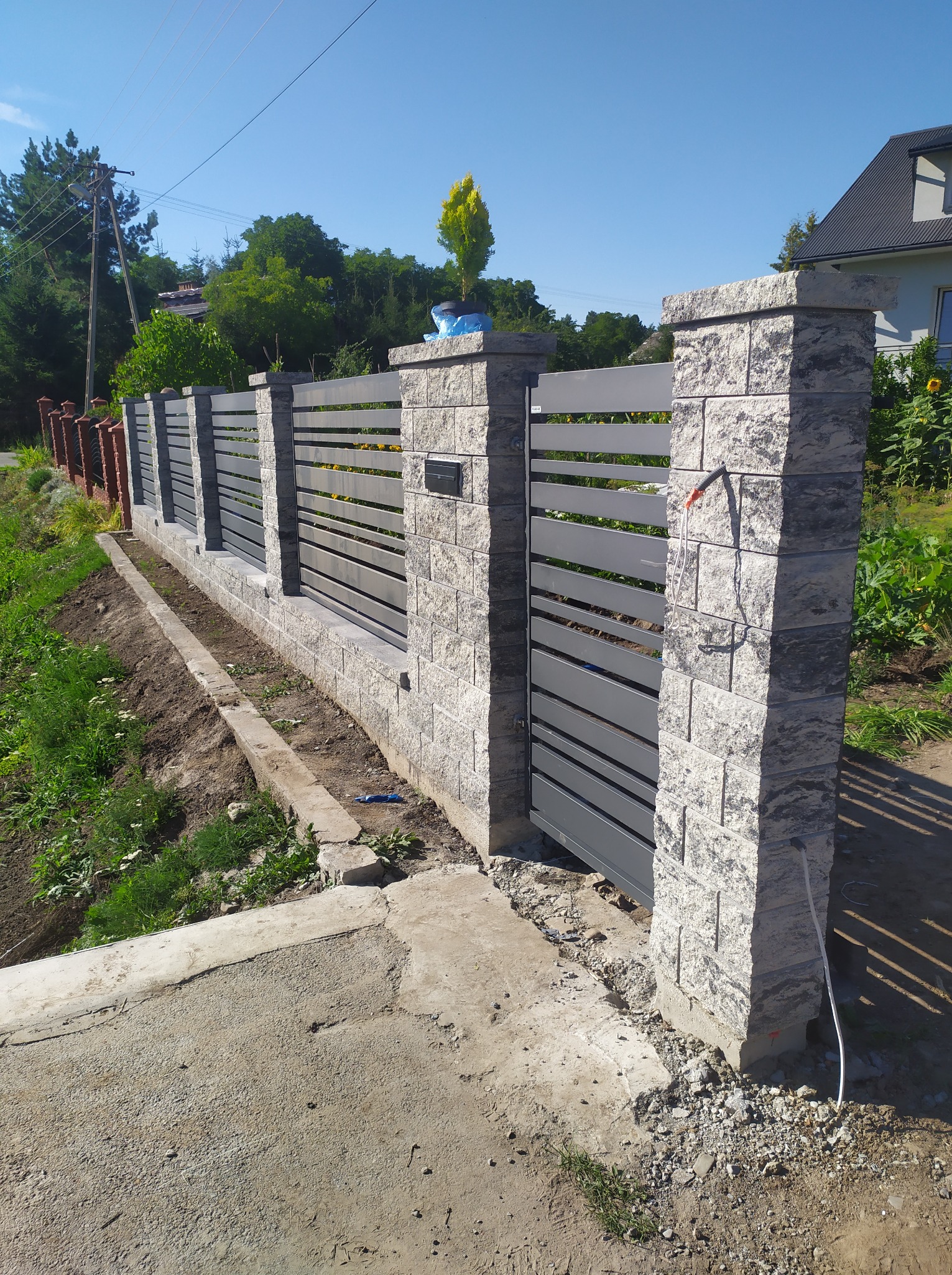Nowoczesne ogrodzenie posesji z szarymi przęsłami i kamiennymi słupkami, świeżo obsadzona ziemia wzdłuż ogrodzenia, brama wjazdowa.