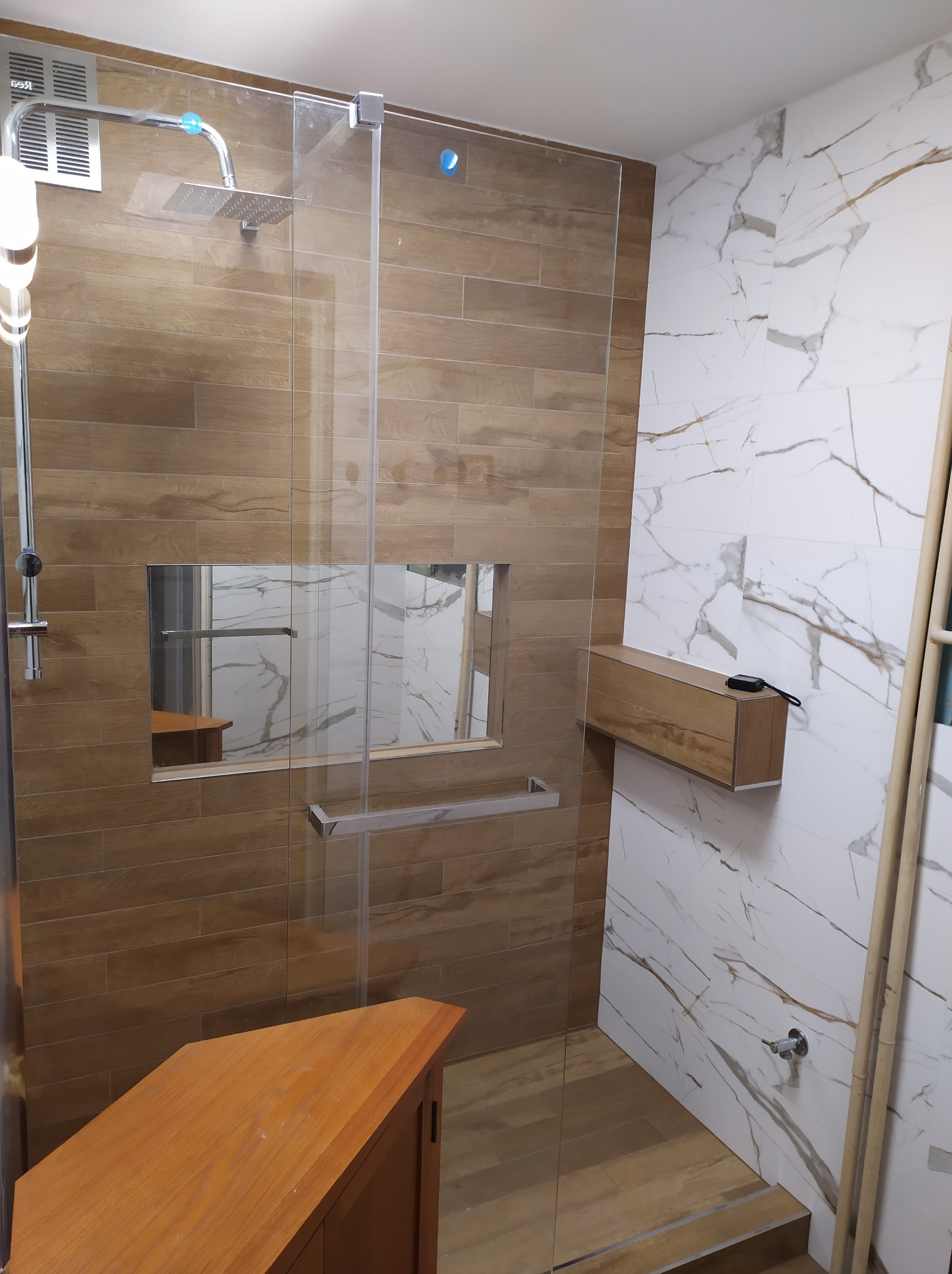 Nowoczesna kabina prysznicowa typu walk-in z drewnopodobnymi płytkami, wbudowaną półką wewnątrz i marmurowymi płytkami na sąsiedniej ścianie, uzupełniona o drewnianą szafkę pod umywalkę i dodatkową...