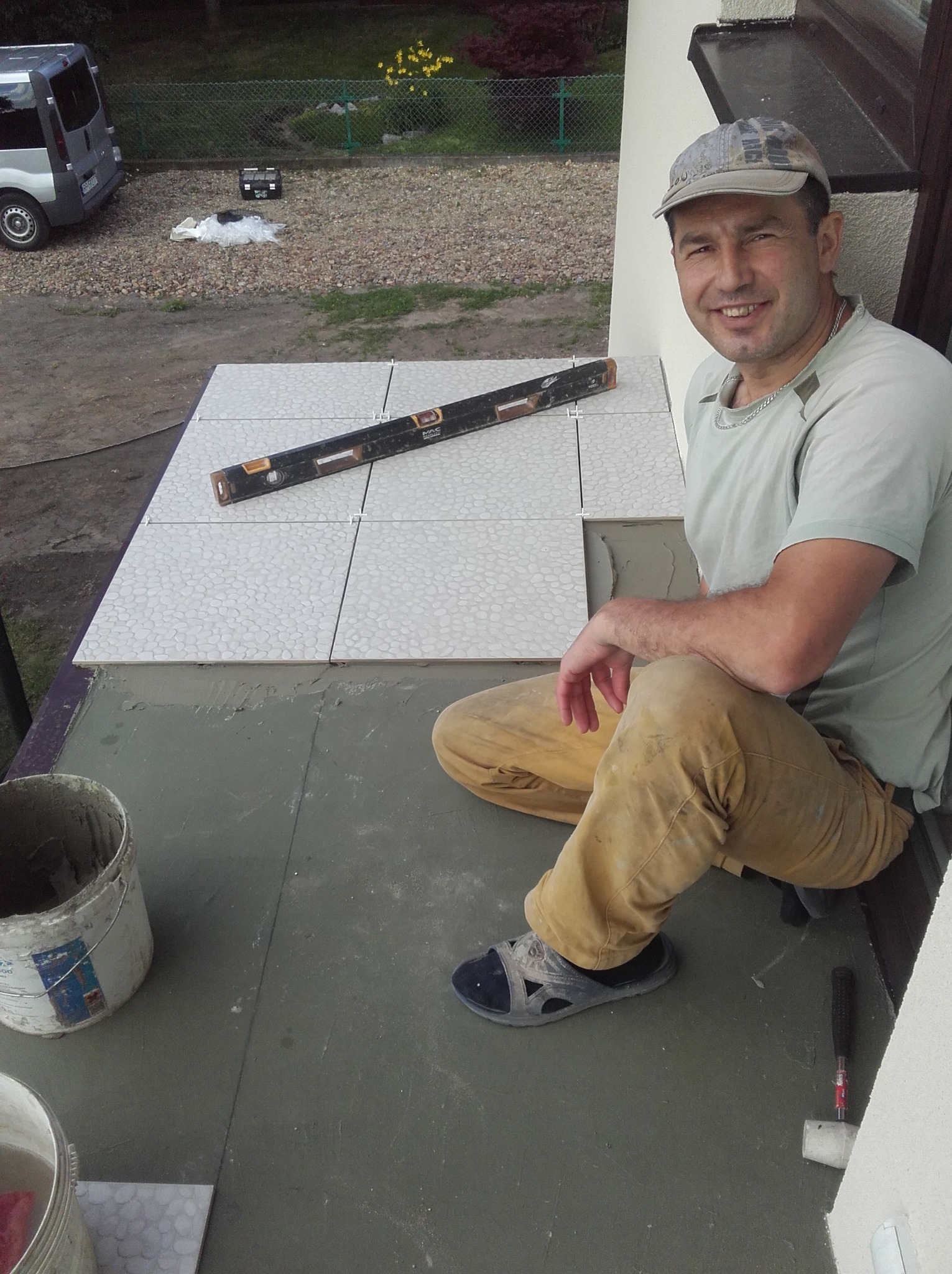 Uśmiechnięty mężczyzna w czapce i roboczych spodniach siedzi na balkonie podczas układania jasnych płytek z wzorem, obok poziomica, wiadro z zaprawą i gumowy młotek.