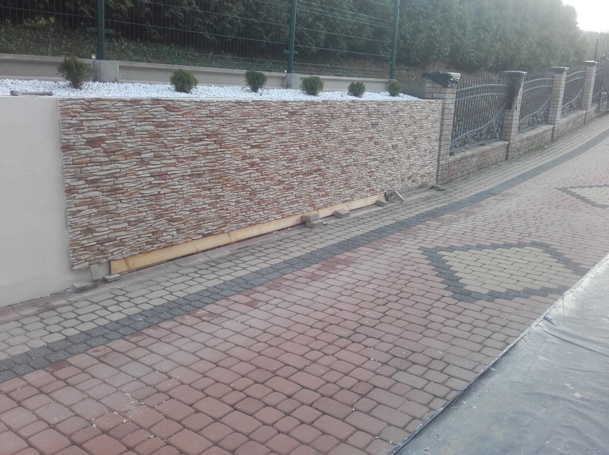Wykończony murek z okładziną imitującą kamień, zwieńczony białymi kamykami i niskimi krzewami, przylegający do kostki brukowej i ozdobnego ogrodzenia.