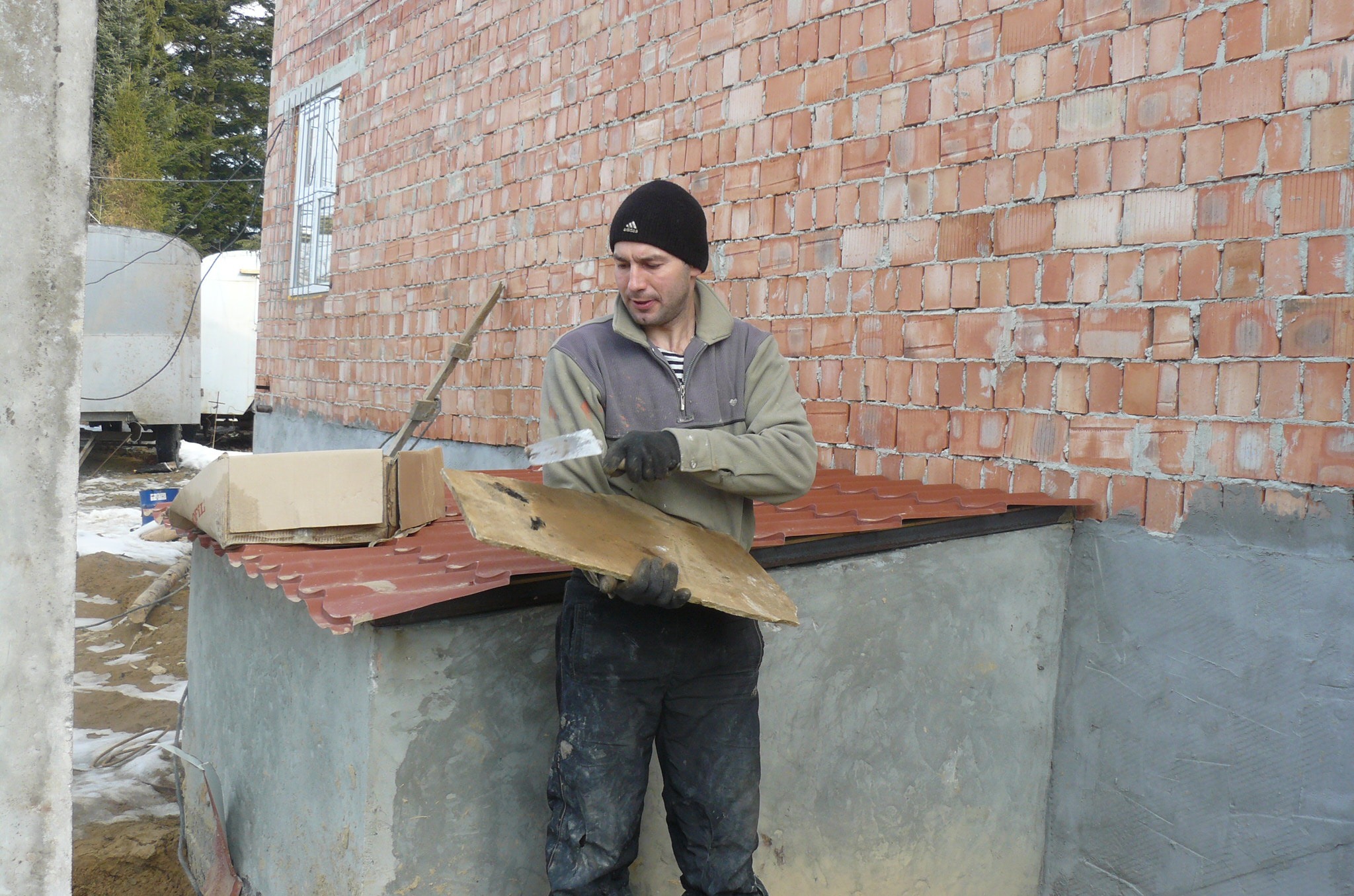 Pracownik w czapce i roboczych ubraniach przygotowuje płytę do montażu, trzymając ją w rękawicach i używając szpachelki, na tle murowanej ściany budynku w trakcie budowy.