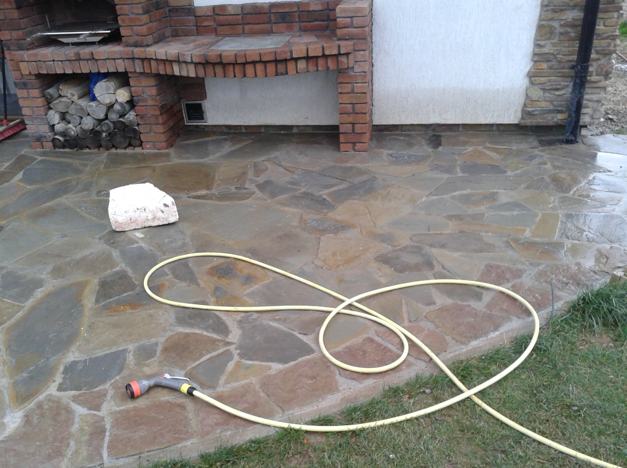Kamienny taras z nieregularnych płyt, mokry po umyciu, z wężem ogrodowym i ceglanym grillem z zapasem drewna w tle.