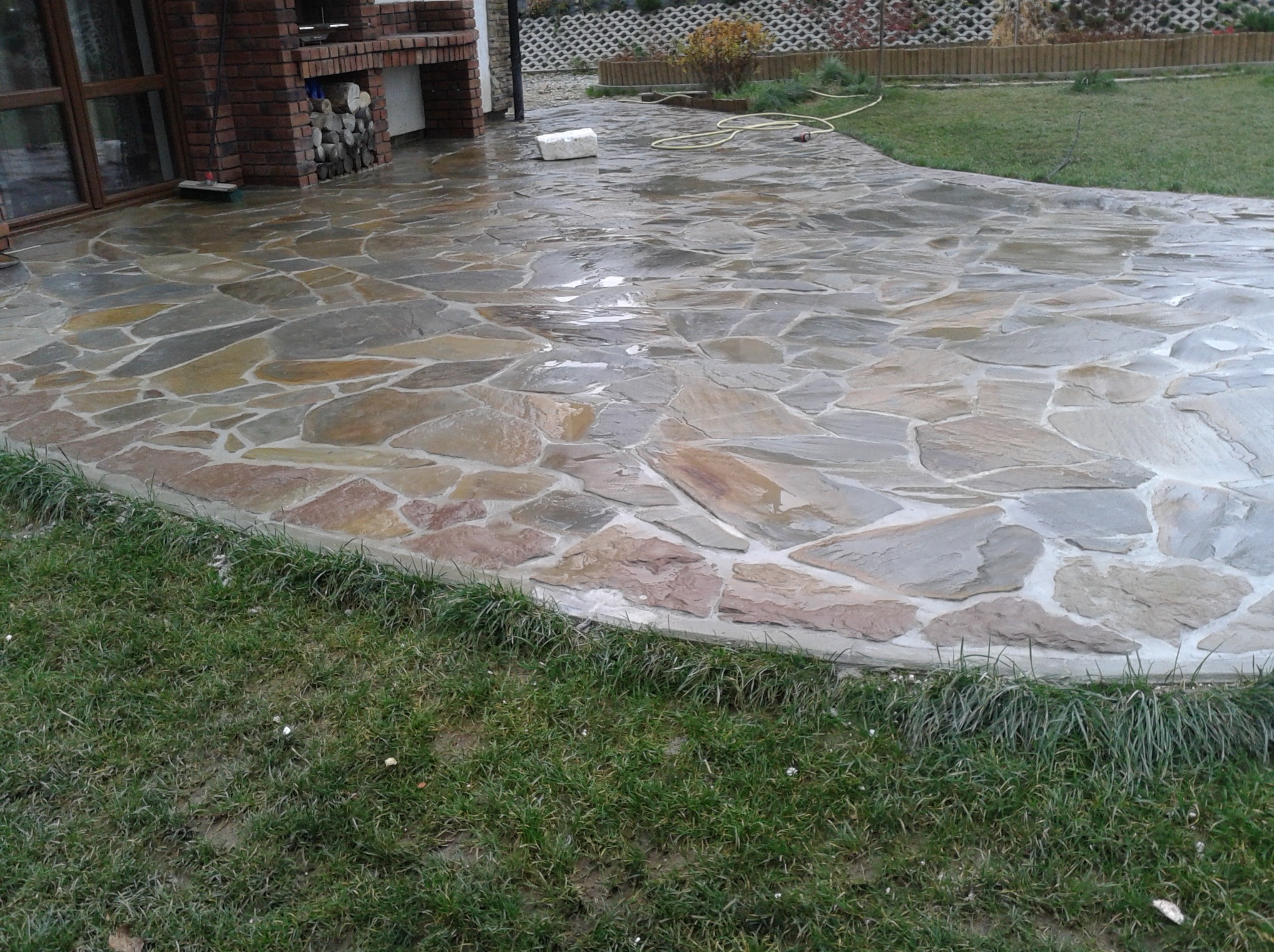 Ułożona z nieregularnych kamieni ścieżka na zewnątrz, obok trawnika i ceglanego grilla, po deszczu.