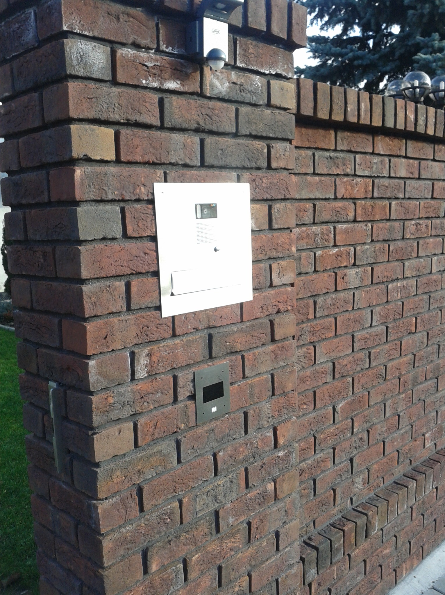 Murowany z cegły klinkierowej słupek ogrodzeniowy z zamontowanym domofonem i oświetleniem zewnętrznym, widoczne ślady użytkowania na cegłach.