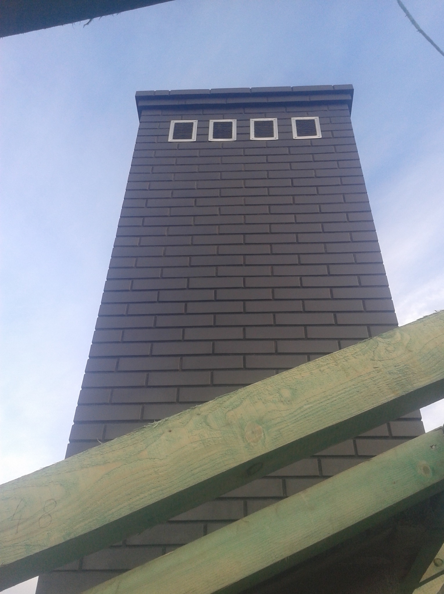 Murowany komin z cegły klinkierowej w kolorze grafitowym z trzema kratkami wentylacyjnymi u góry, widoczny z dołu, częściowo zasłonięty przez zielone krokwie dachowe.