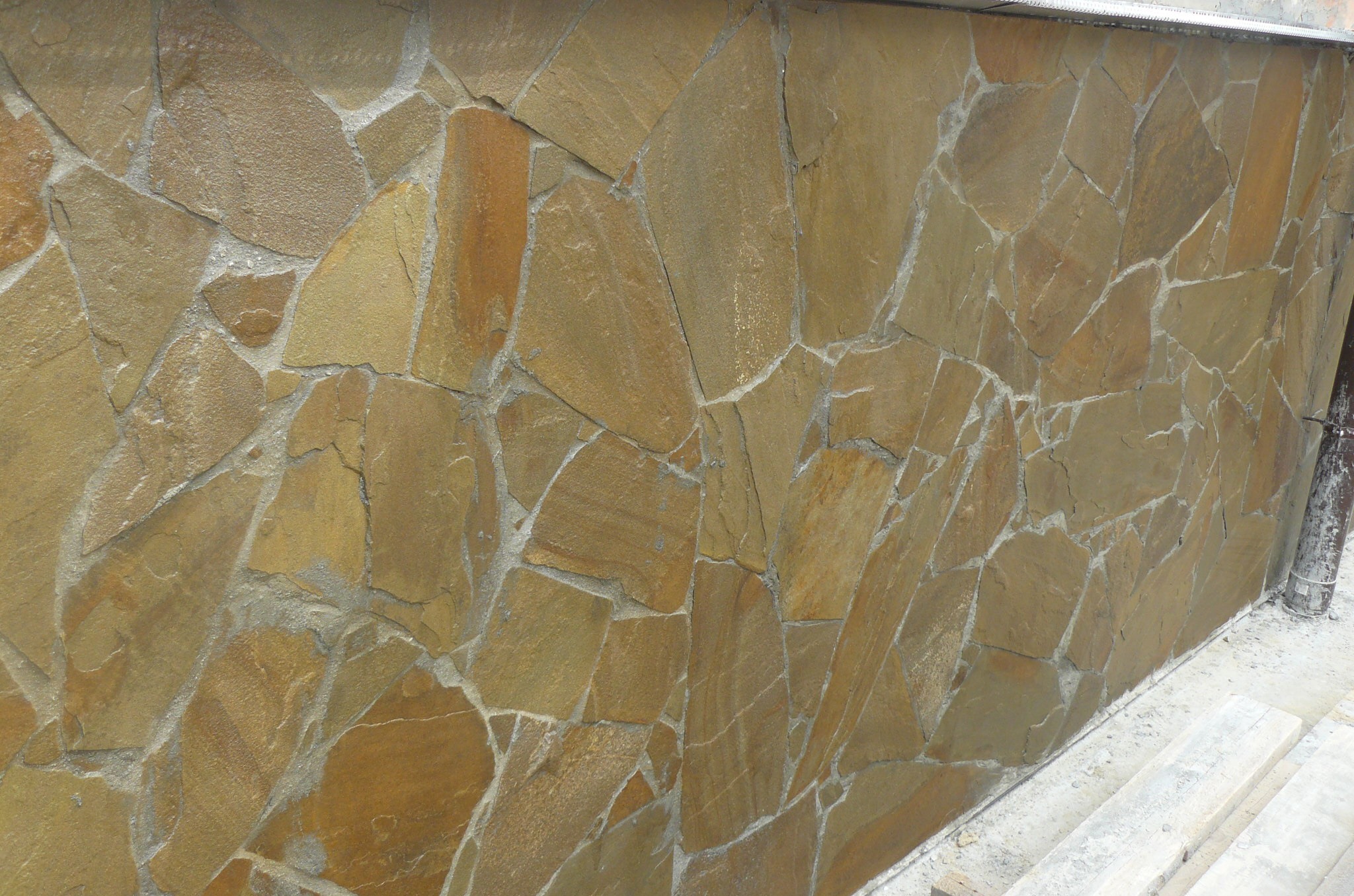 Ściana wykończona nieregularnymi płytami z piaskowca w odcieniach beżu i brązu, z widocznymi spoinami wypełnionymi szarą zaprawą, przy podstawie widoczny fragment posadzki i rury spustowej.