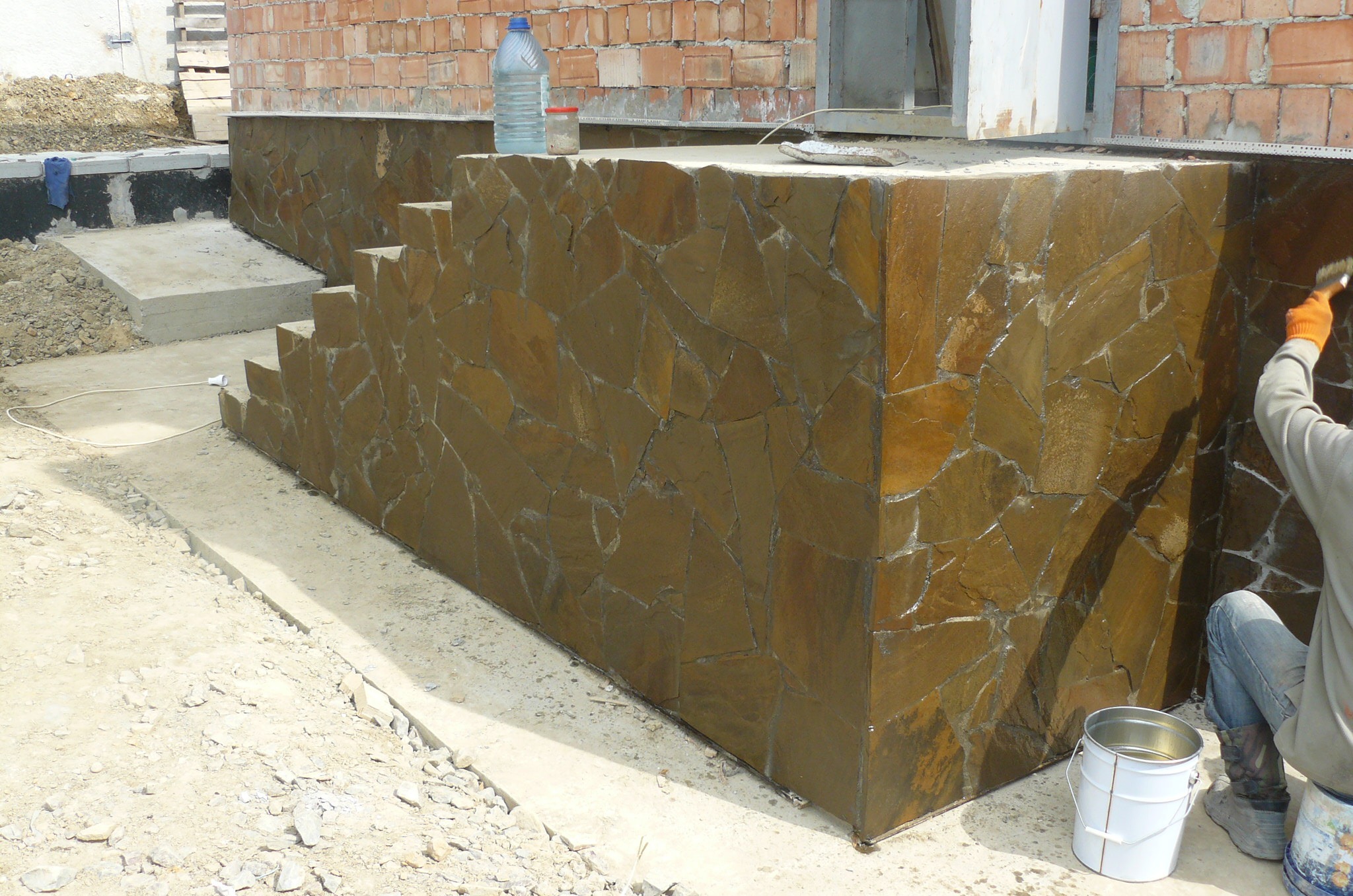 Wykończenie elewacji budynku kamieniem naturalnym w odcieniach brązu, widoczne schody wejściowe i pracownik impregnujący kamień pędzlem, wiadro z impregnatem stoi obok.