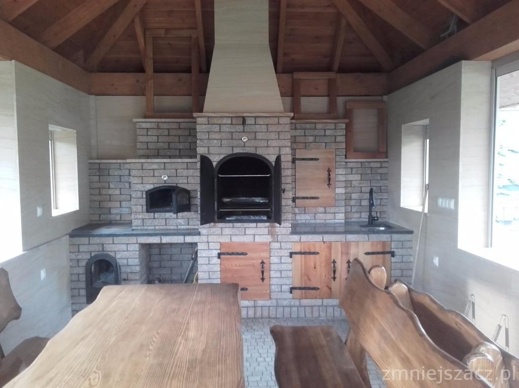 Zadaszona kuchnia letnia z murowanym grillem, piekarnikiem i blatem roboczym z szarej cegły oraz drewnianymi szafkami i meblami w rustykalnym stylu.