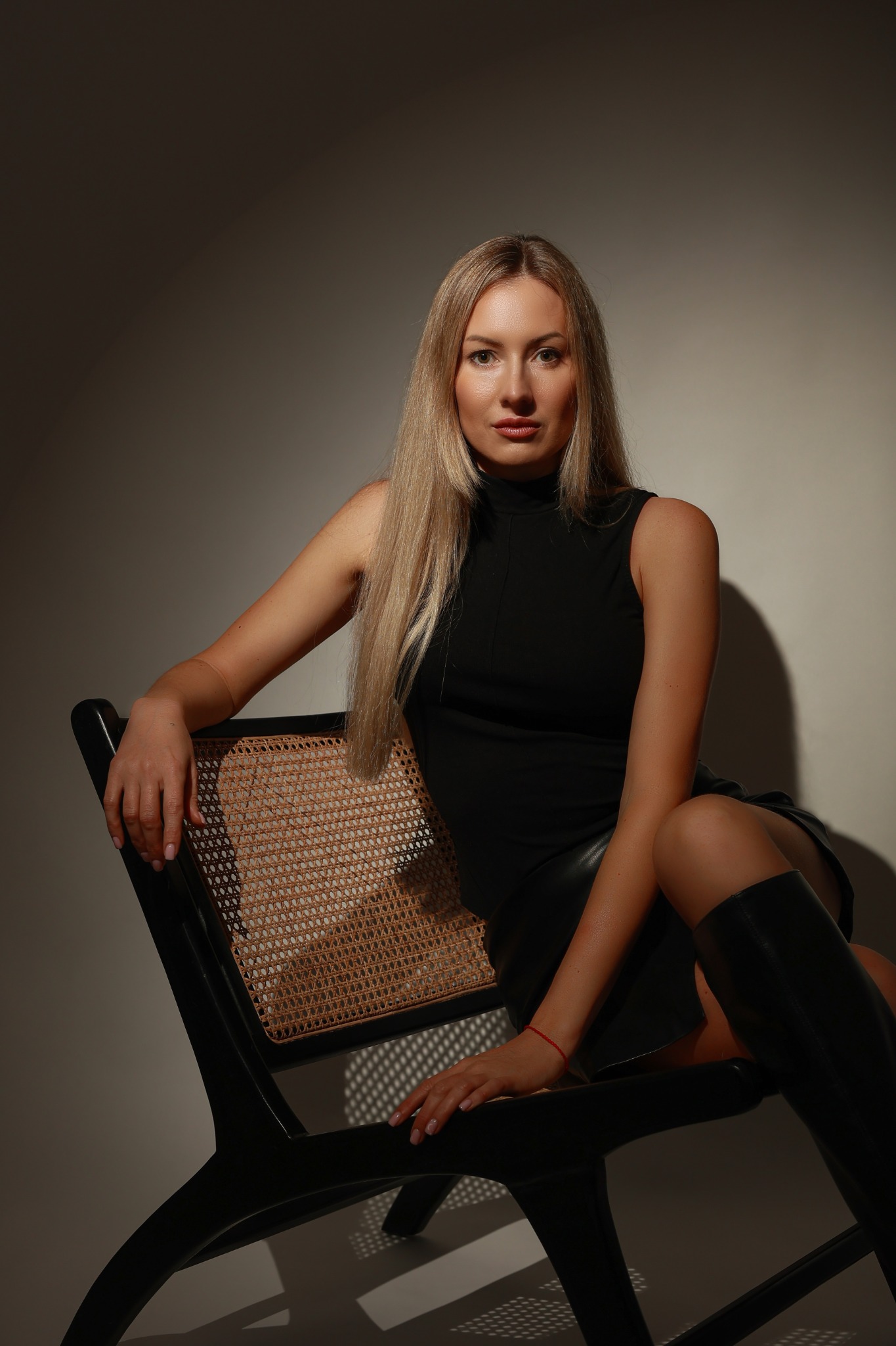 Portret kobiety z długimi blond włosami, ubranej w czarną sukienkę i kozaki, siedzącej na designerskim krześle z plecionym oparciem, oświetlonej punktowym światłem.