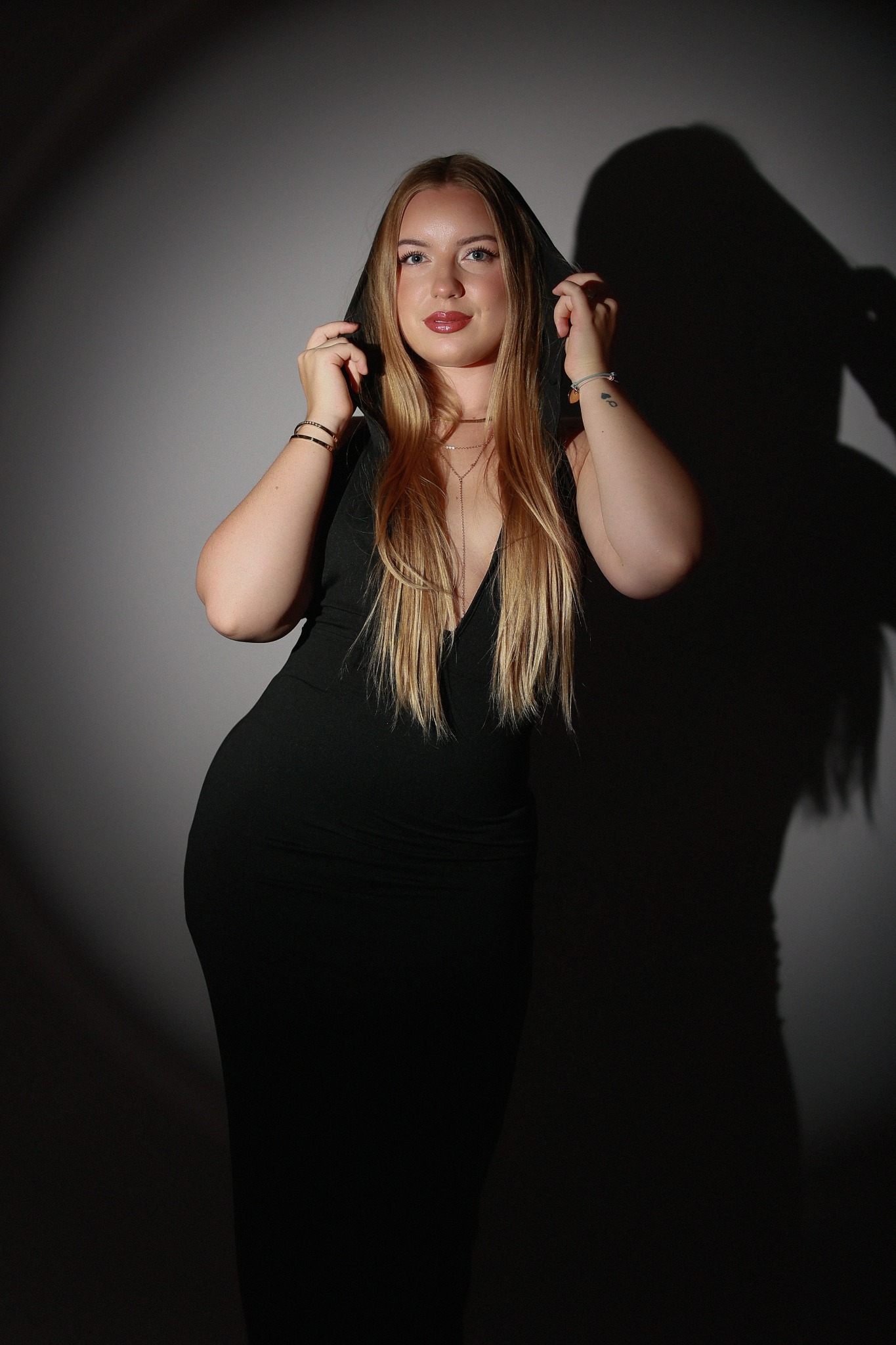 Portret kobiety w czarnej sukience z kapturem, oświetlonej punktowo, z widocznym cieniem na szarym tle.