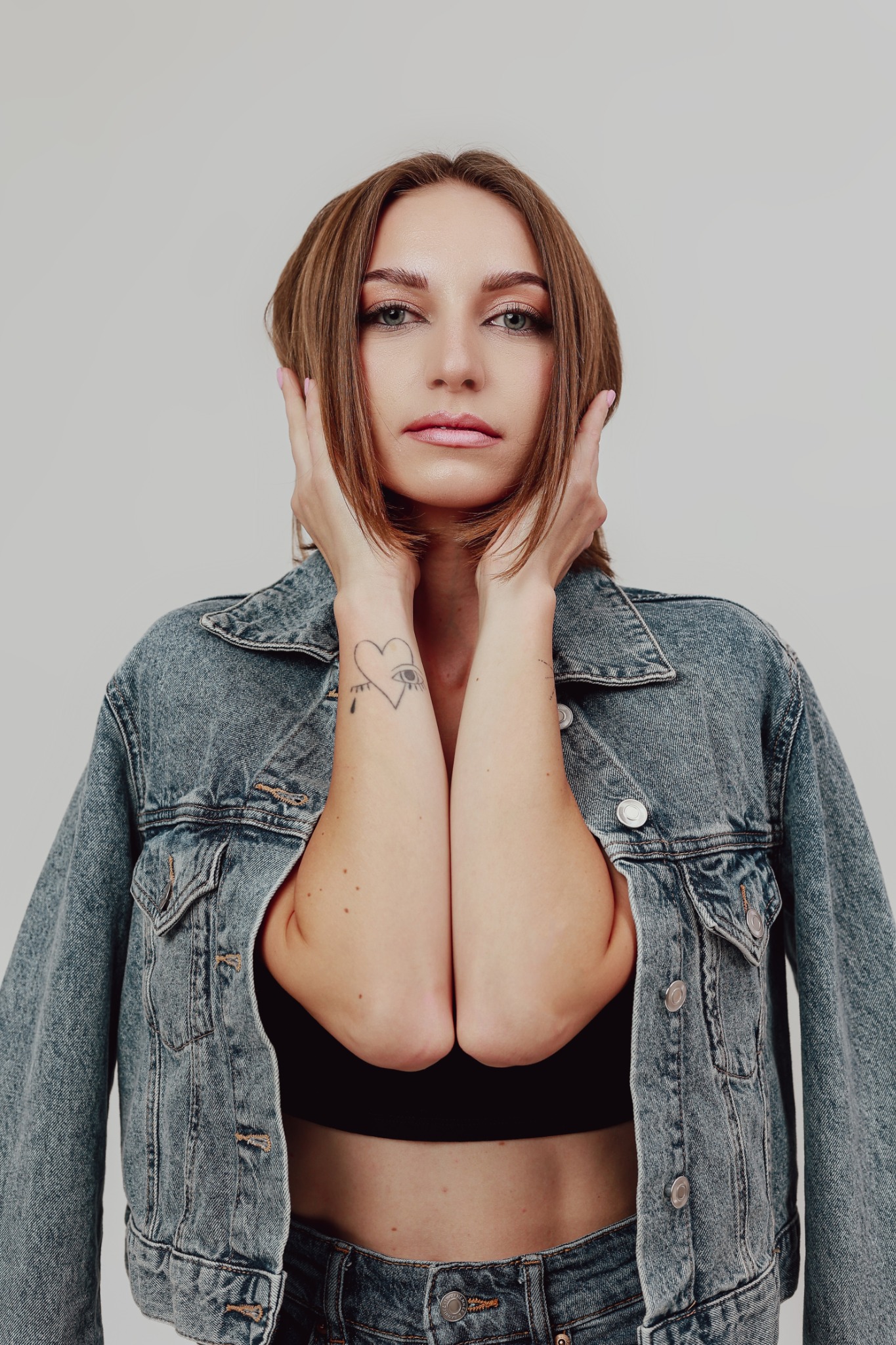 Portret kobiety w dżinsowej kurtce, z rękami przy twarzy, ukazujący tatuaż serca z okiem na przedramieniu, na jasnym tle.