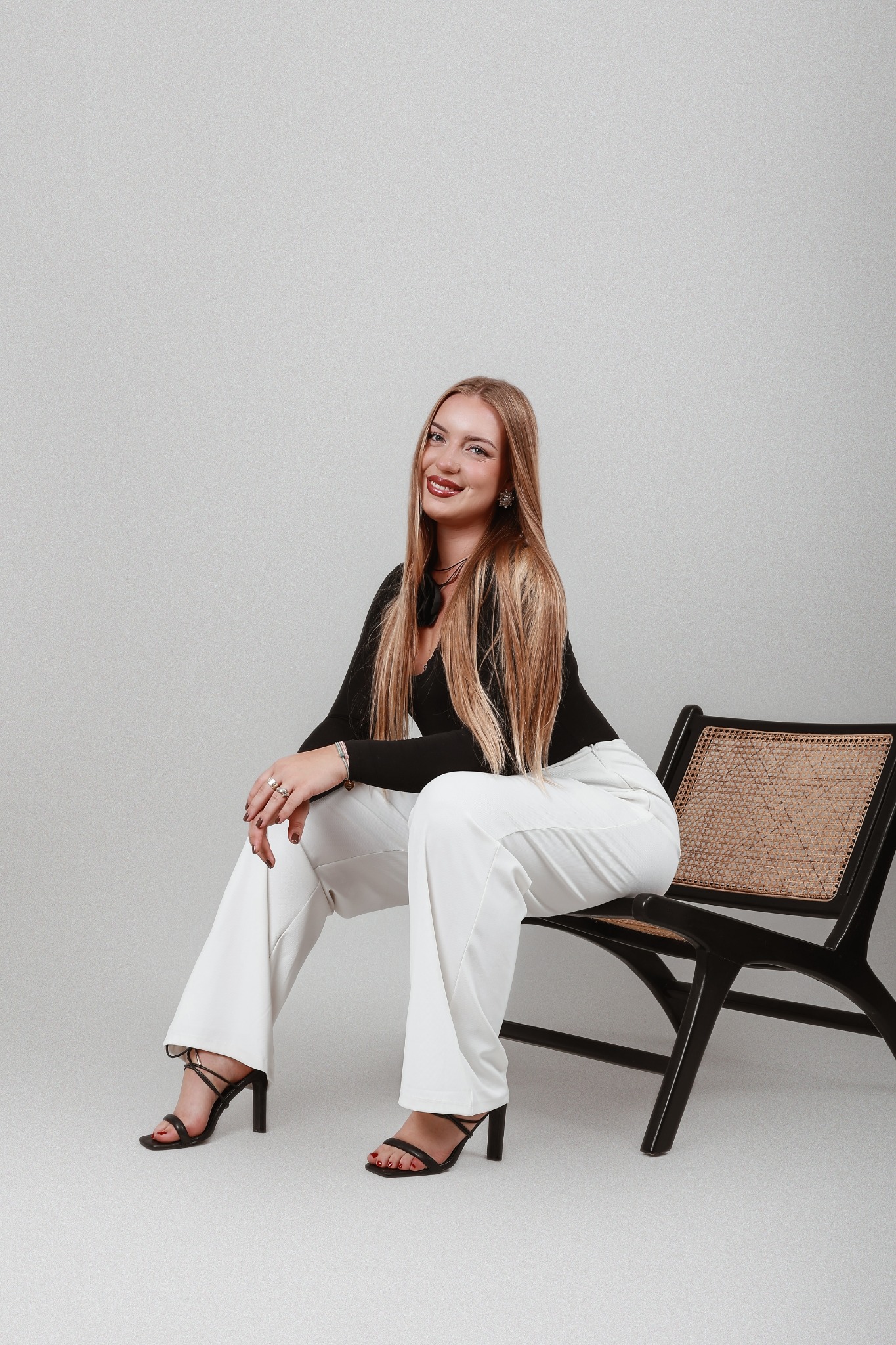 Portret uśmiechniętej, długowłosej blondynki w czarnej bluzce i białych spodniach, siedzącej na czarnym krześle z rattanowym siedziskiem, na jasnoszarym tle.